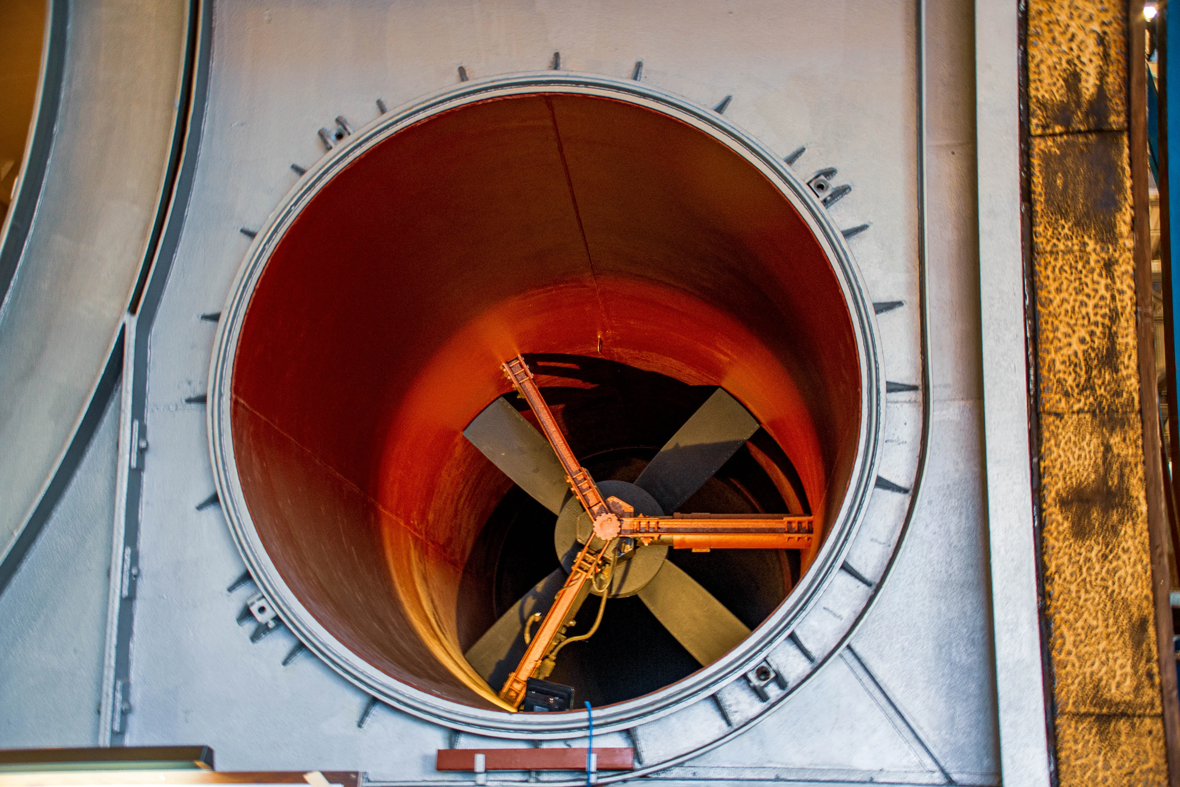 Close-up of a large, complex industrial wind tunnel. A wide, circular aperture dominates the centre, divided into four sections, each leading to a smaller circular opening. Surrounding this are large pipes, green industrial machinery, and metallic structural elements.