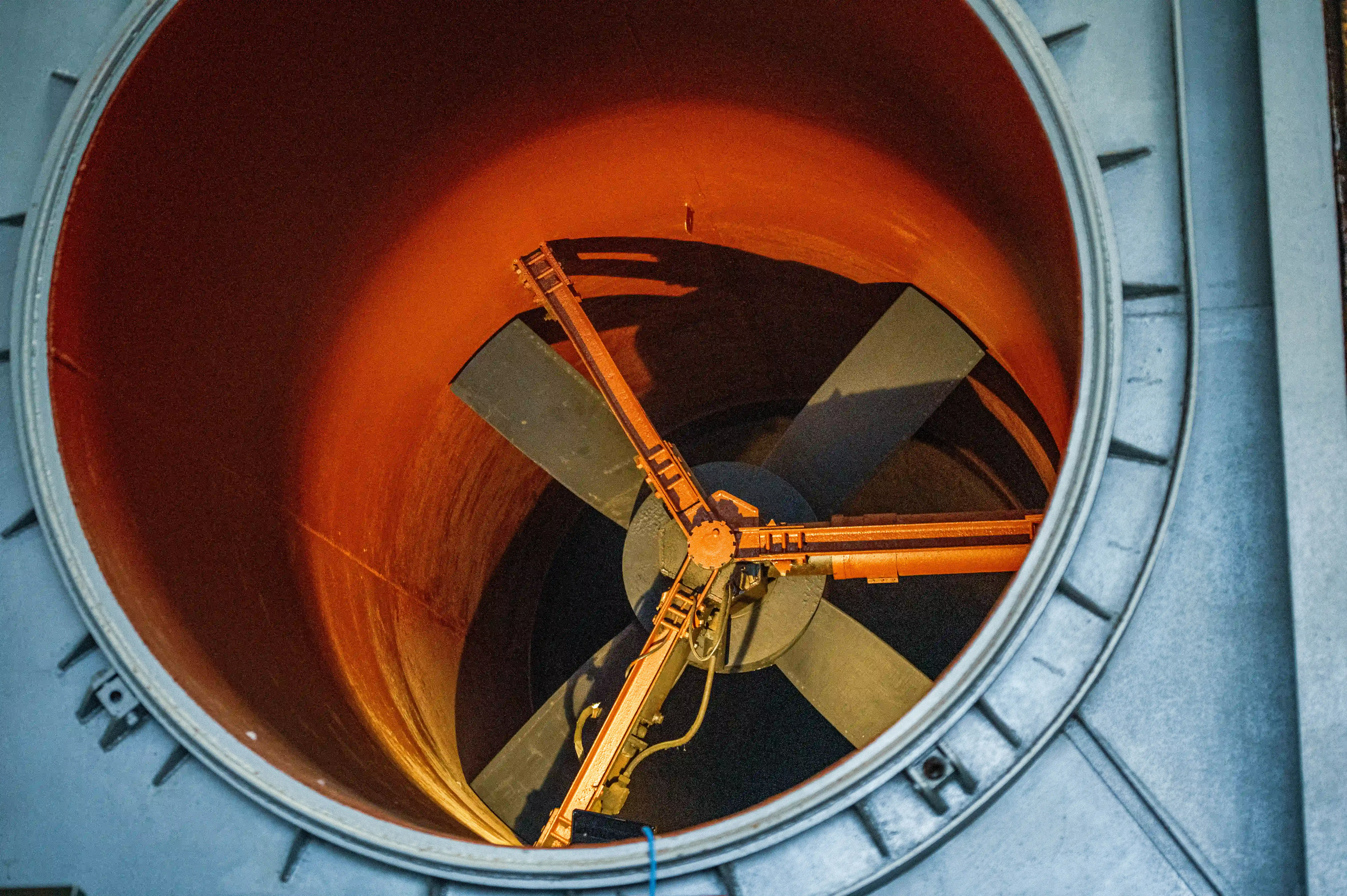 Close-up of a large, complex industrial wind tunnel. A wide, circular aperture dominates the centre, divided into four sections, each leading to a smaller circular opening. Surrounding this are large pipes, green industrial machinery, and metallic structural elements.