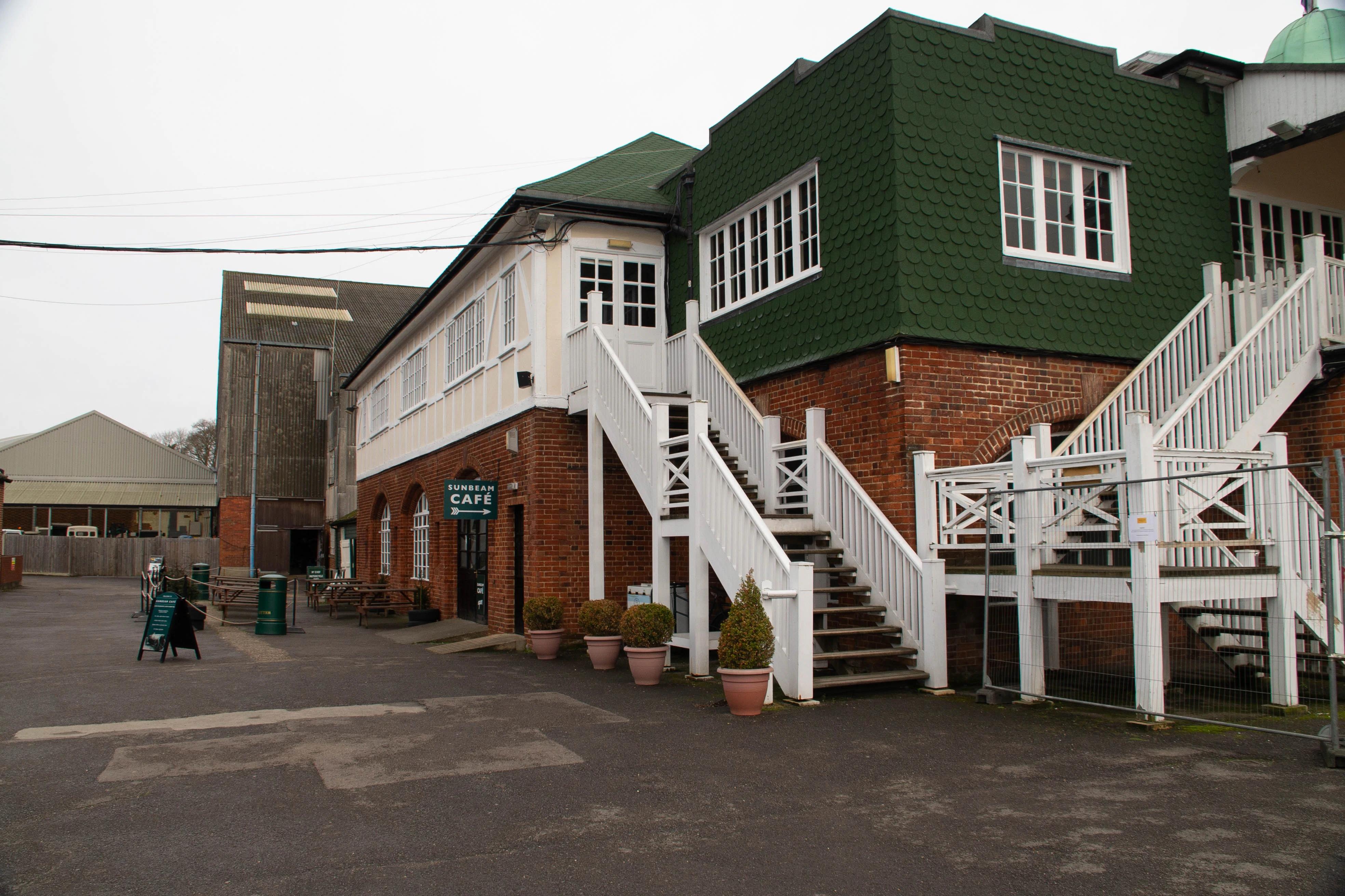 A two-storey building with a green shingled roof and red brick lower section. A white wooden staircase with balustrades runs up the side of the building. A sign indicates SUNBEAM CAFE. In the background, a tall, dark industrial building is visible.