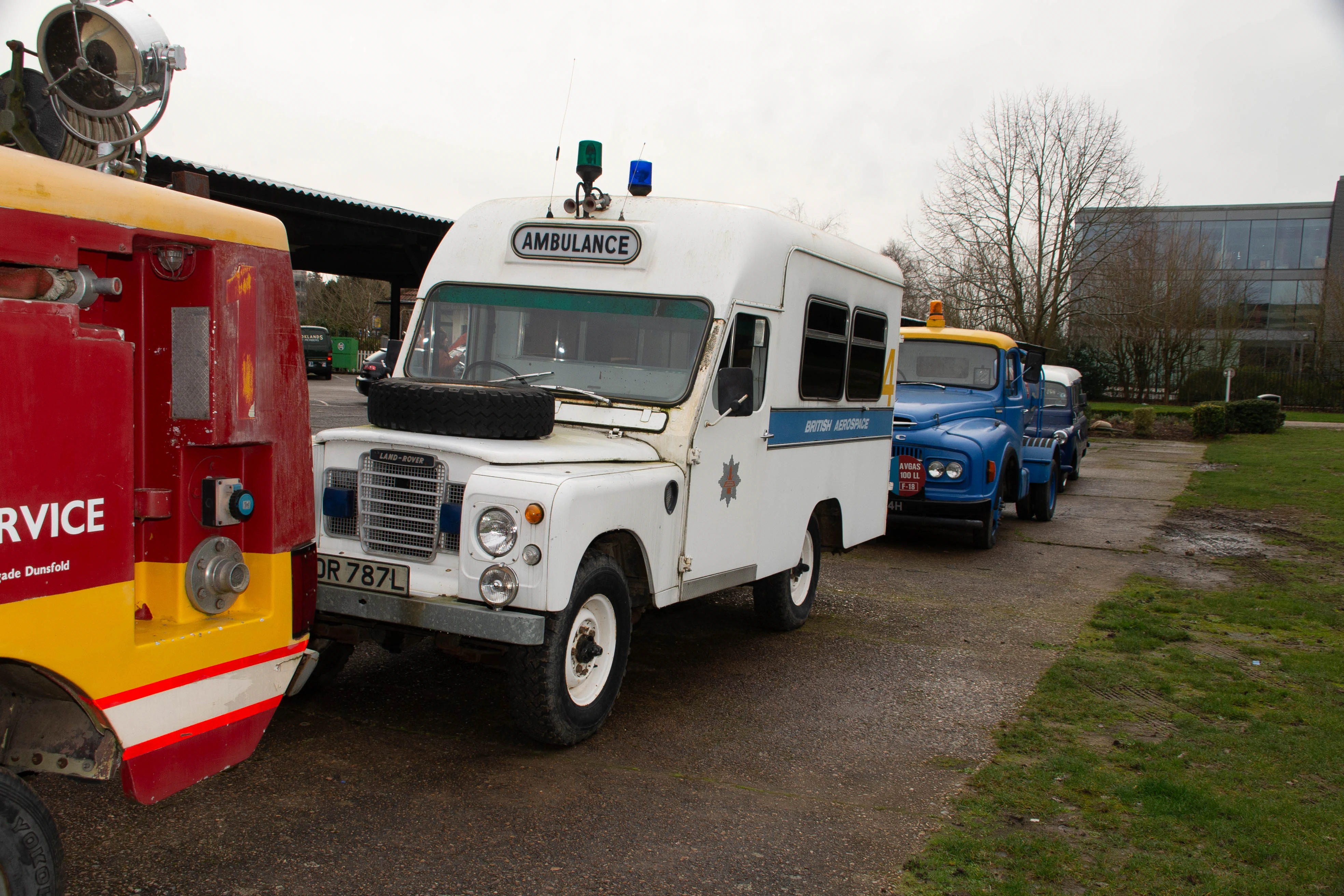 A white vintage Land Rover ambulance with blue and green flashing lights is parked on a tarmac surface. Beside it is the rear of a red and yellow emergency vehicle. In the background, several blue trucks are lined up.