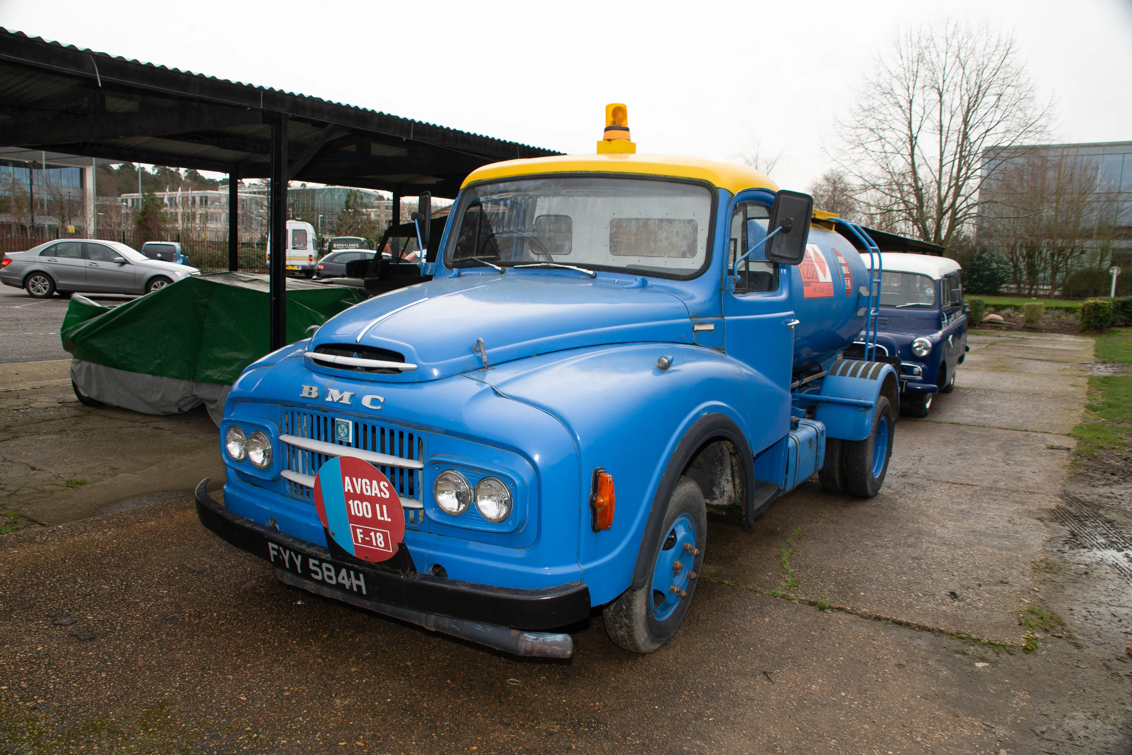 A classic blue BMC tanker truck with a yellow roof and an orange flashing beacon is parked on a wet concrete lot. A round decal on the front of the truck reads AVGAS 100 LL F-18. A vintage blue Land Rover is parked behind the truck. Modern buildings and trees are visible in the background.