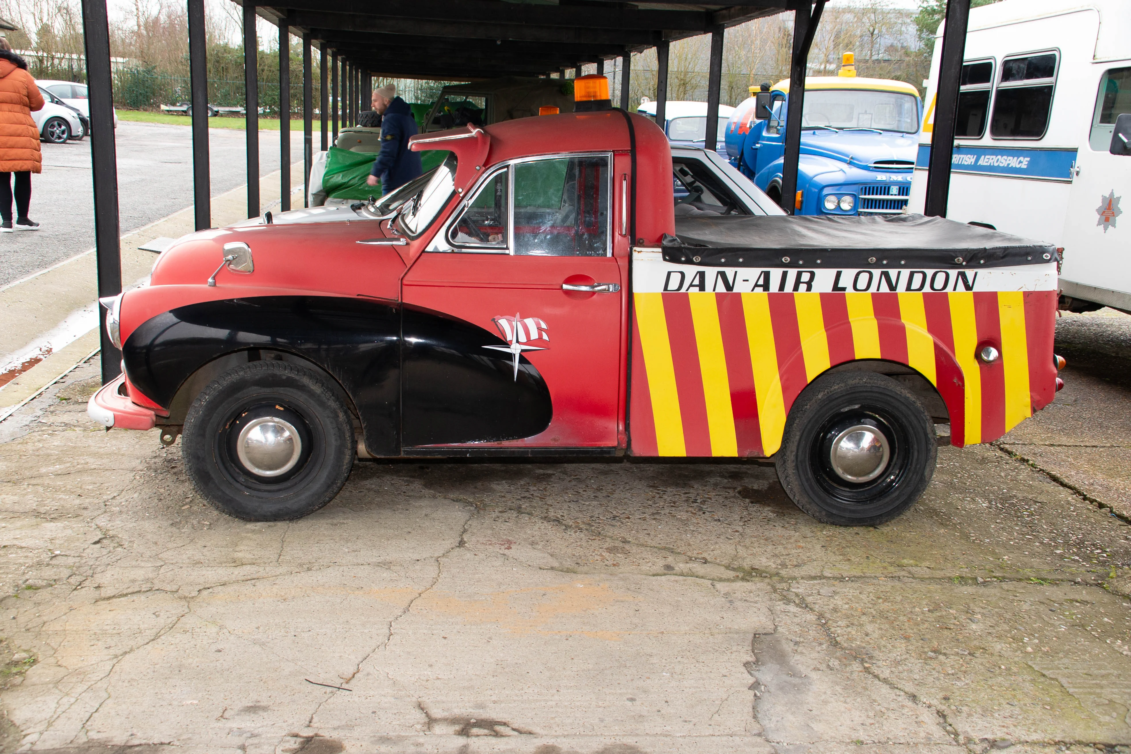 Side view of a vintage red and black van with Dan-Air London written on the back in white lettering. The rear of the van has bold red and yellow diagonal stripes. It has chrome hubcaps and a chrome wing mirror. Other vehicles, including a blue truck with 