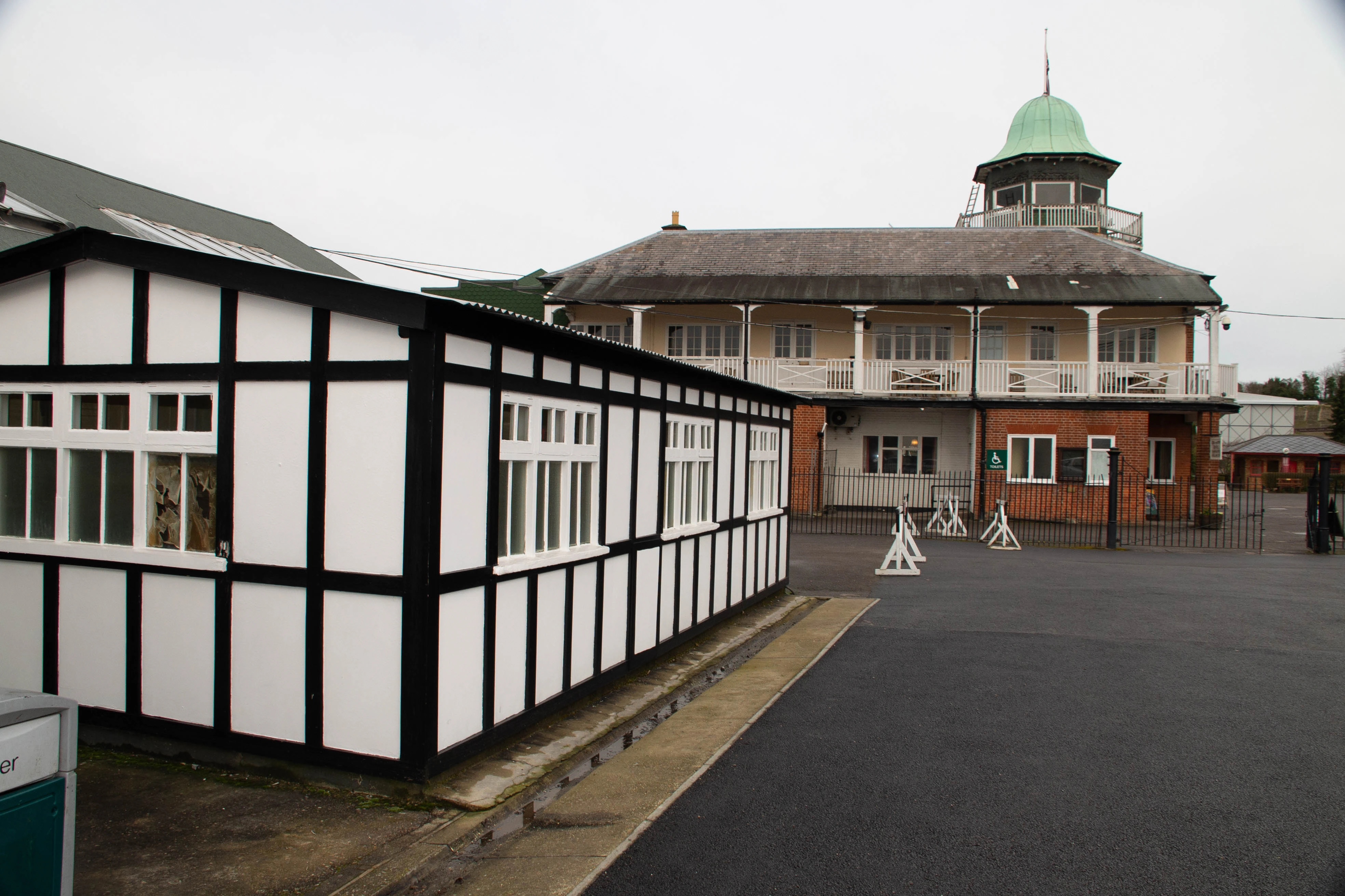 A black and white timber-framed building with multiple windows is positioned in the foreground, angled to the left. Behind it stands a larger, multi-story brick building with a yellow facade and white balconies, topped by a prominent green domed cupola. The sky is overcast
