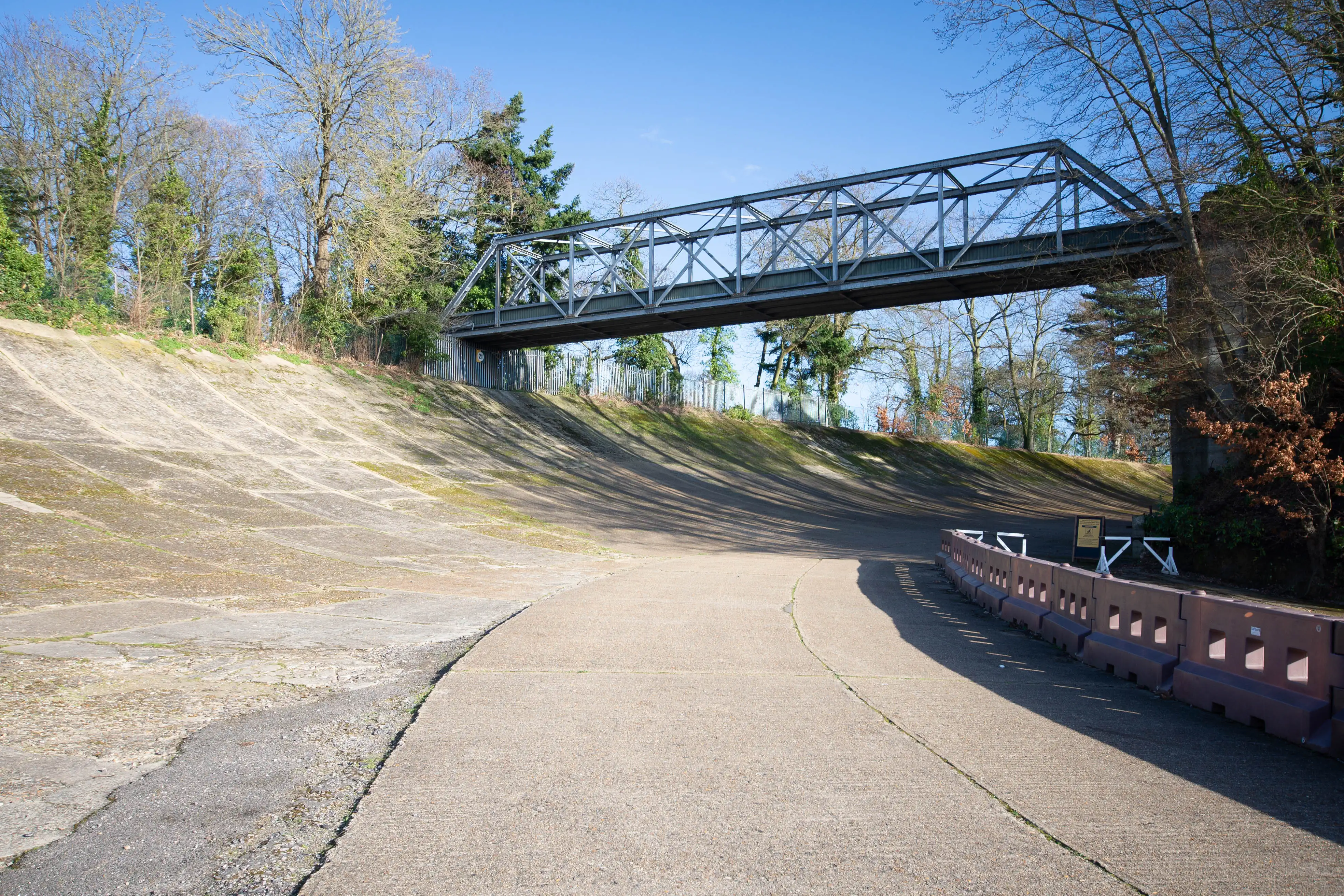 A view of the steeply banked concrete curve of the historic Brooklands race circuit, with a metal bridge overhead and trees in the background under a bright blue sky.