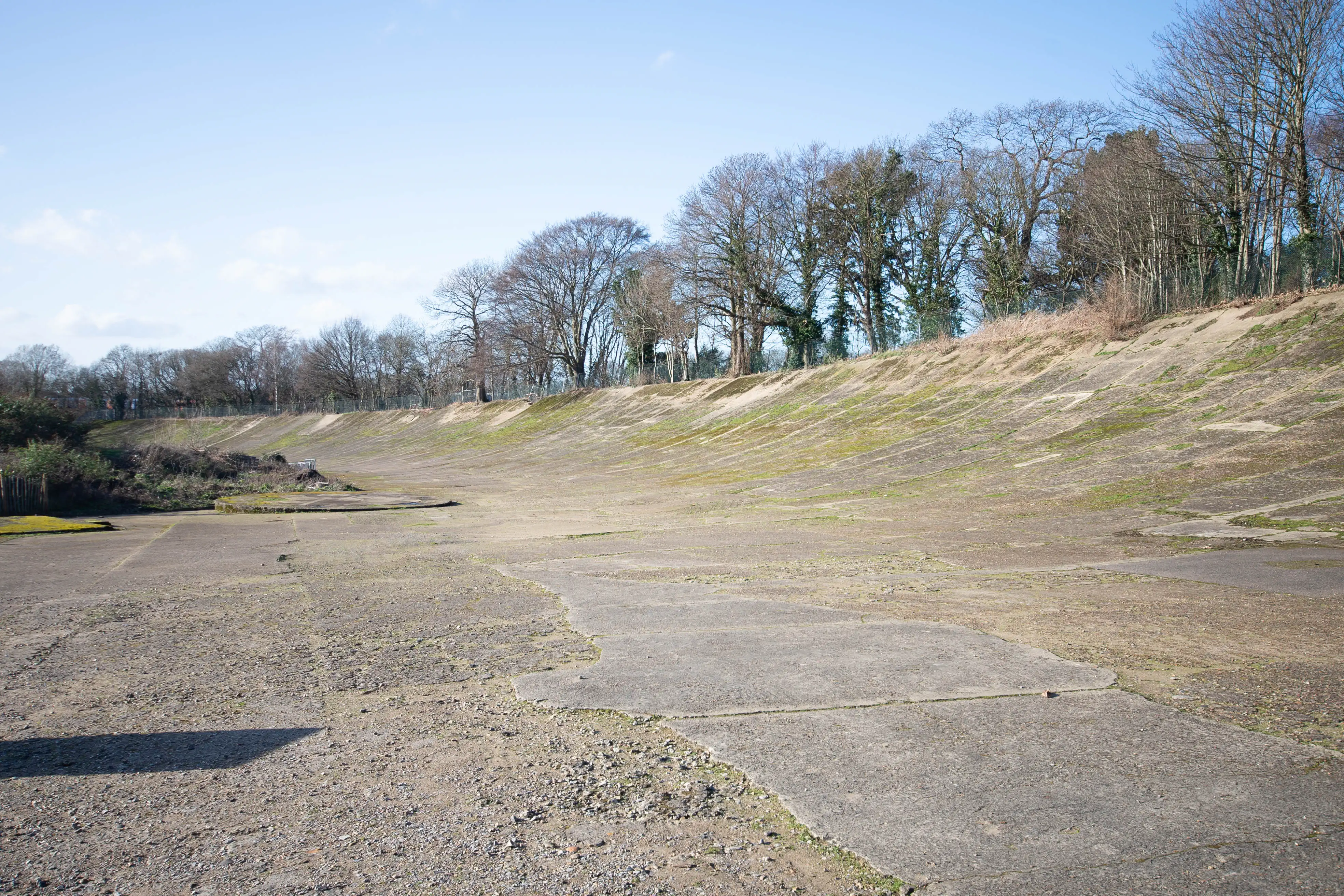 A wide shot of the empty, banked concrete track of the historic Brooklands racing circuit, with grass growing on the slopes and bare trees in the background under a blue sky.