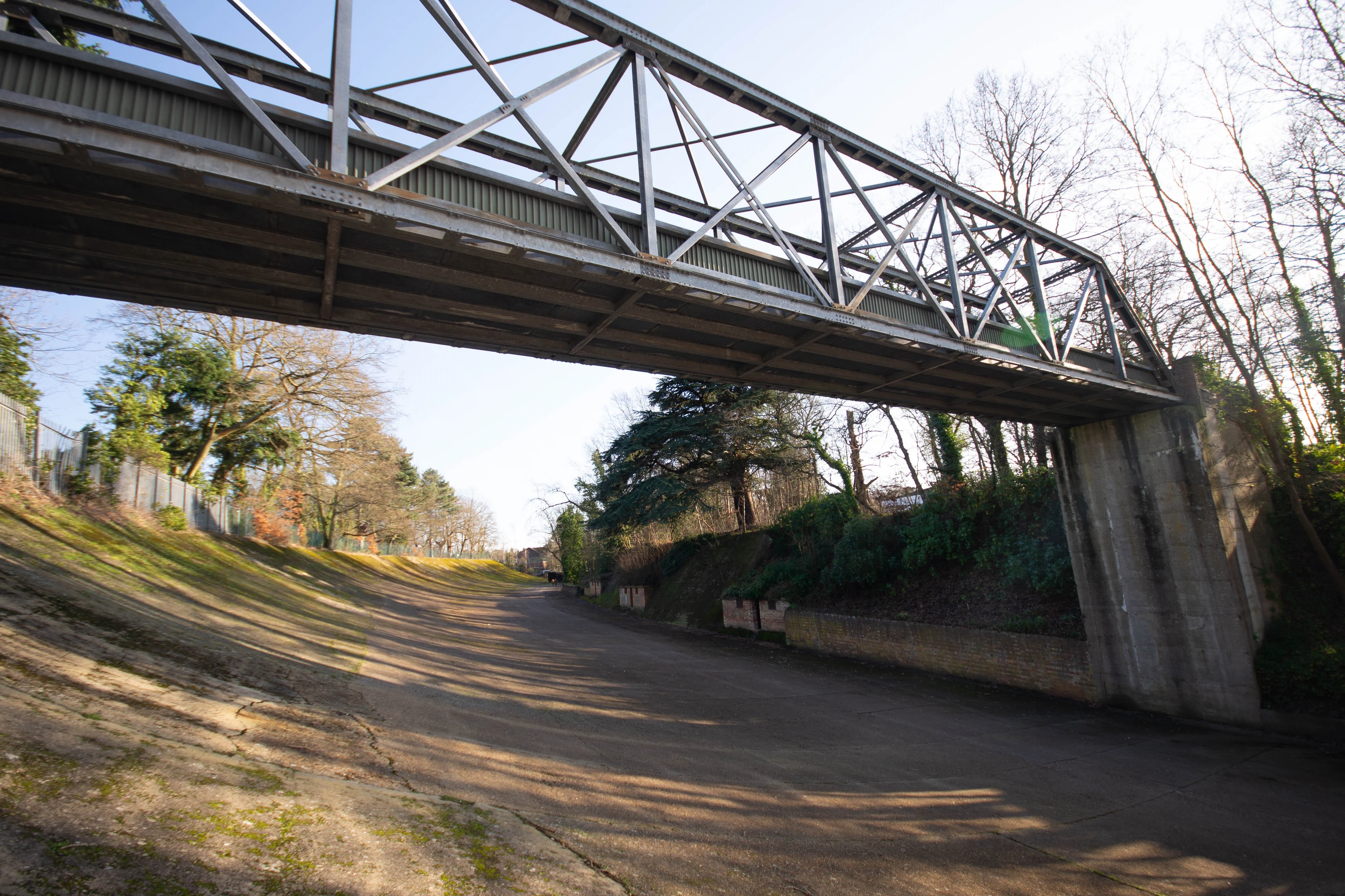 A wide shot of a concrete and grass lined disused railway track, curving to the right and disappearing into the distance. A large metal truss railway bridge spans overhead, supported by concrete pillars. Trees and foliage line the banks, and dappled sunlight casts shadows across the track