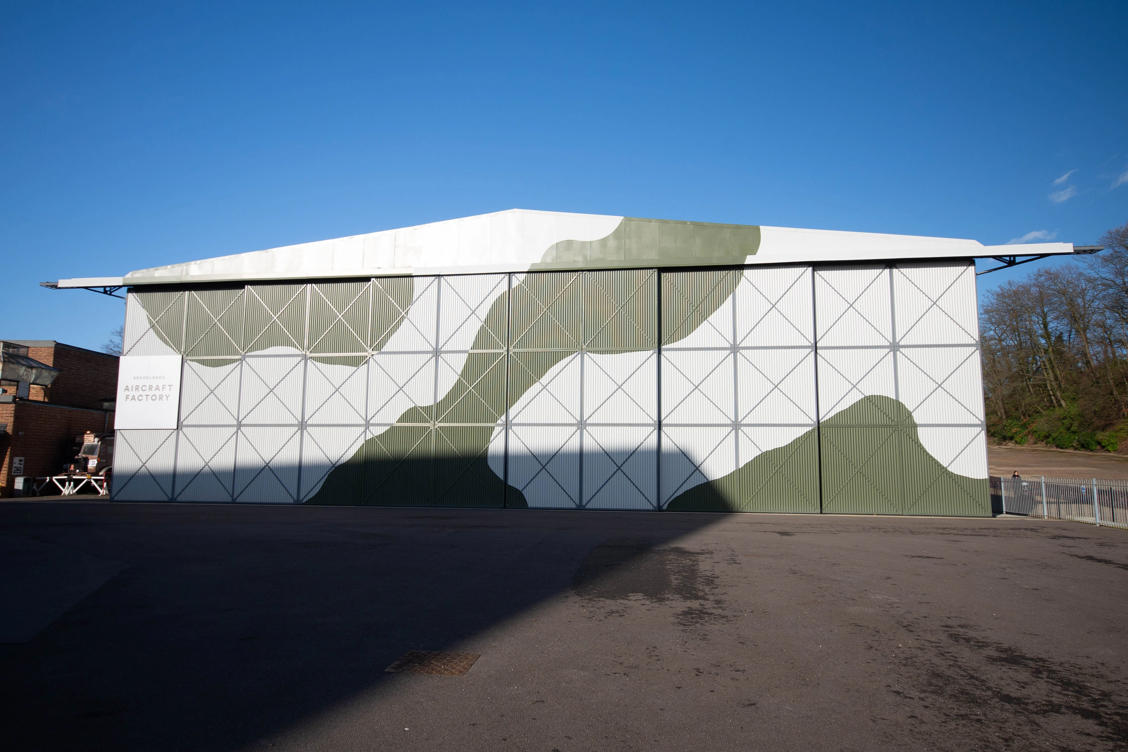 A large, white industrial building with sections of green camouflage painted on its corrugated metal doors. The building has a sign that reads BROOKLANDS AIRCRAFT FACTORY.