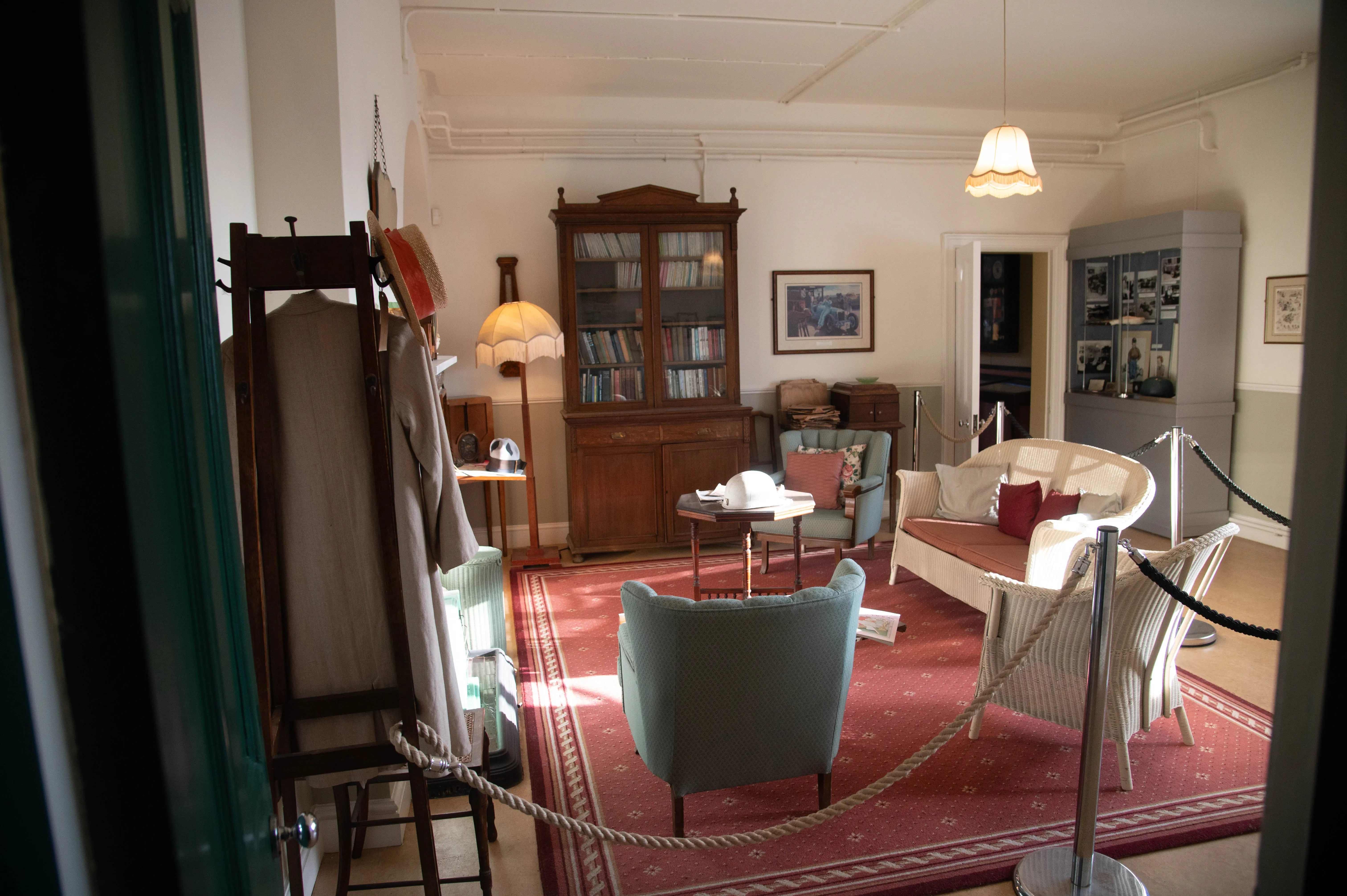 A vintage sitting room furnished with a wooden bookshelf, a light blue armchair, a wicker sofa with red and grey cushions, a red patterned carpet, a coat stand with clothing and hats, and a floor lamp with a fringed shade.