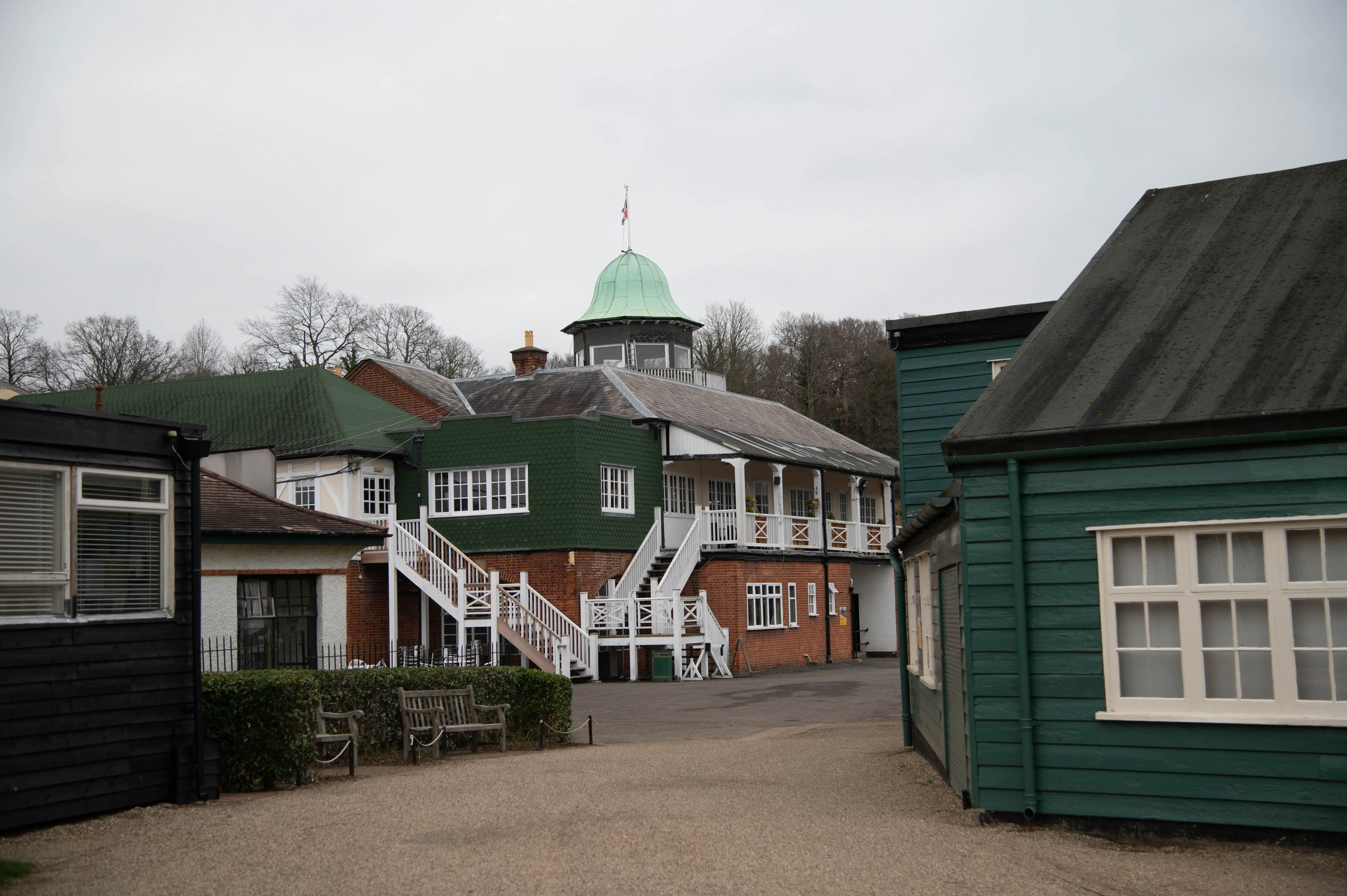 An exterior shot of a vintage cricket club building with a prominent green domed tower topped by a flag. White wooden stairs lead up to a balcony. The building has red brickwork and white balconies with patterned supports. Adjacent are dark grey and bright green wooden structures.