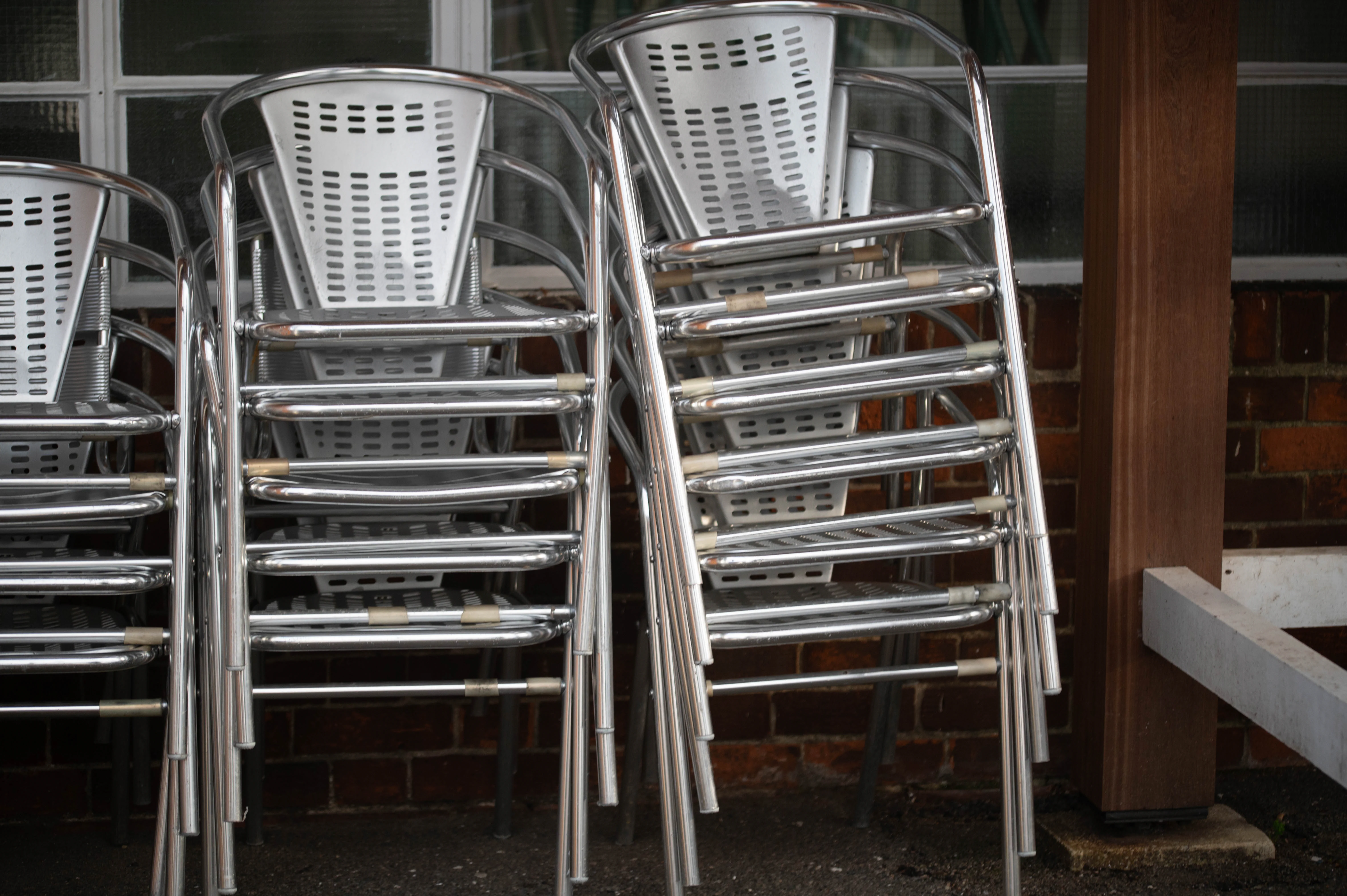 Two stacks of silver metal chairs with perforated backrests placed neatly against a brick wall.