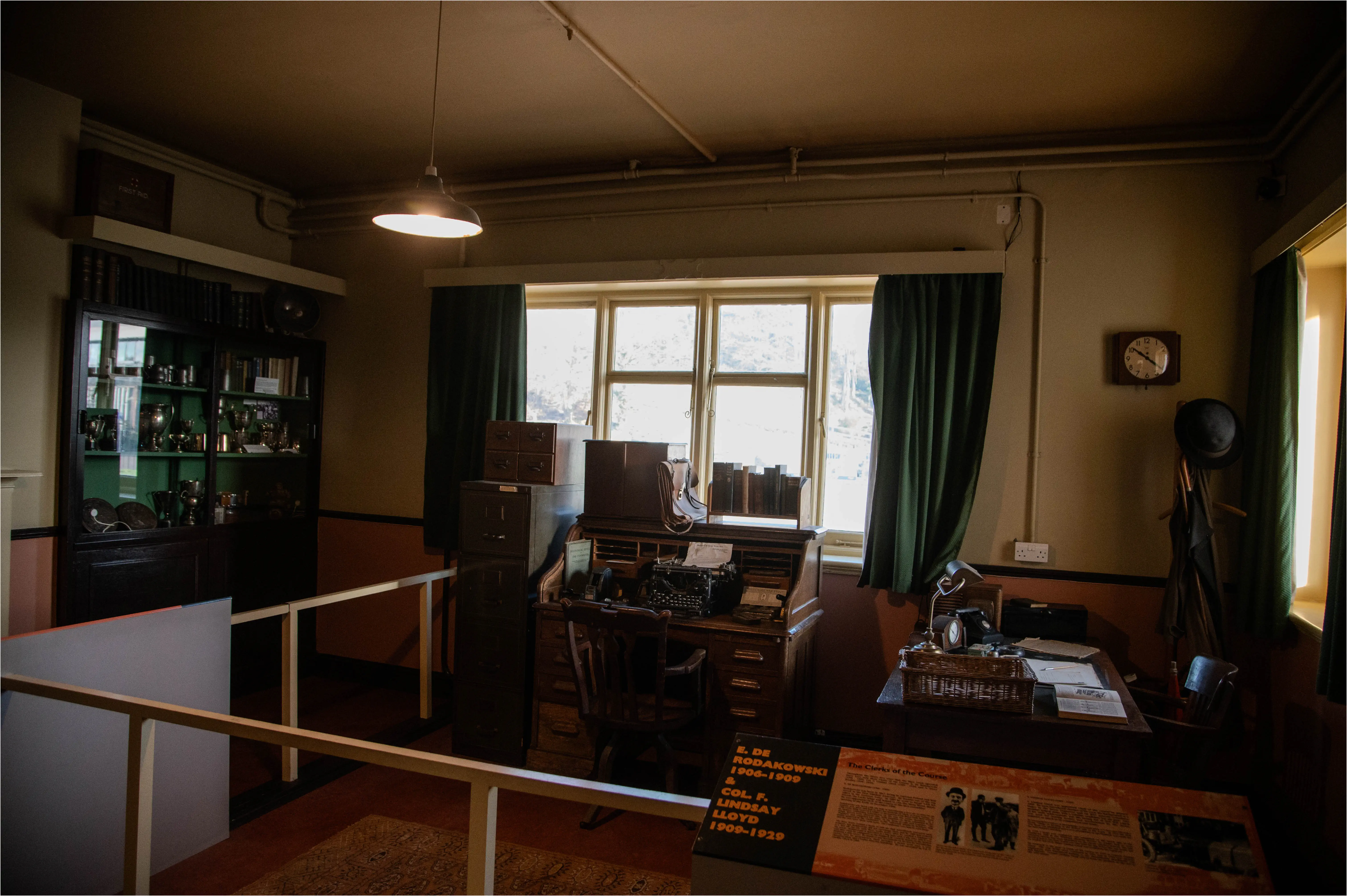 An interior view of a recreated historical office, showing a wooden desk with a typewriter, a chair, filing cabinets, a glass-fronted cabinet filled with trophies, and a window with green curtains.