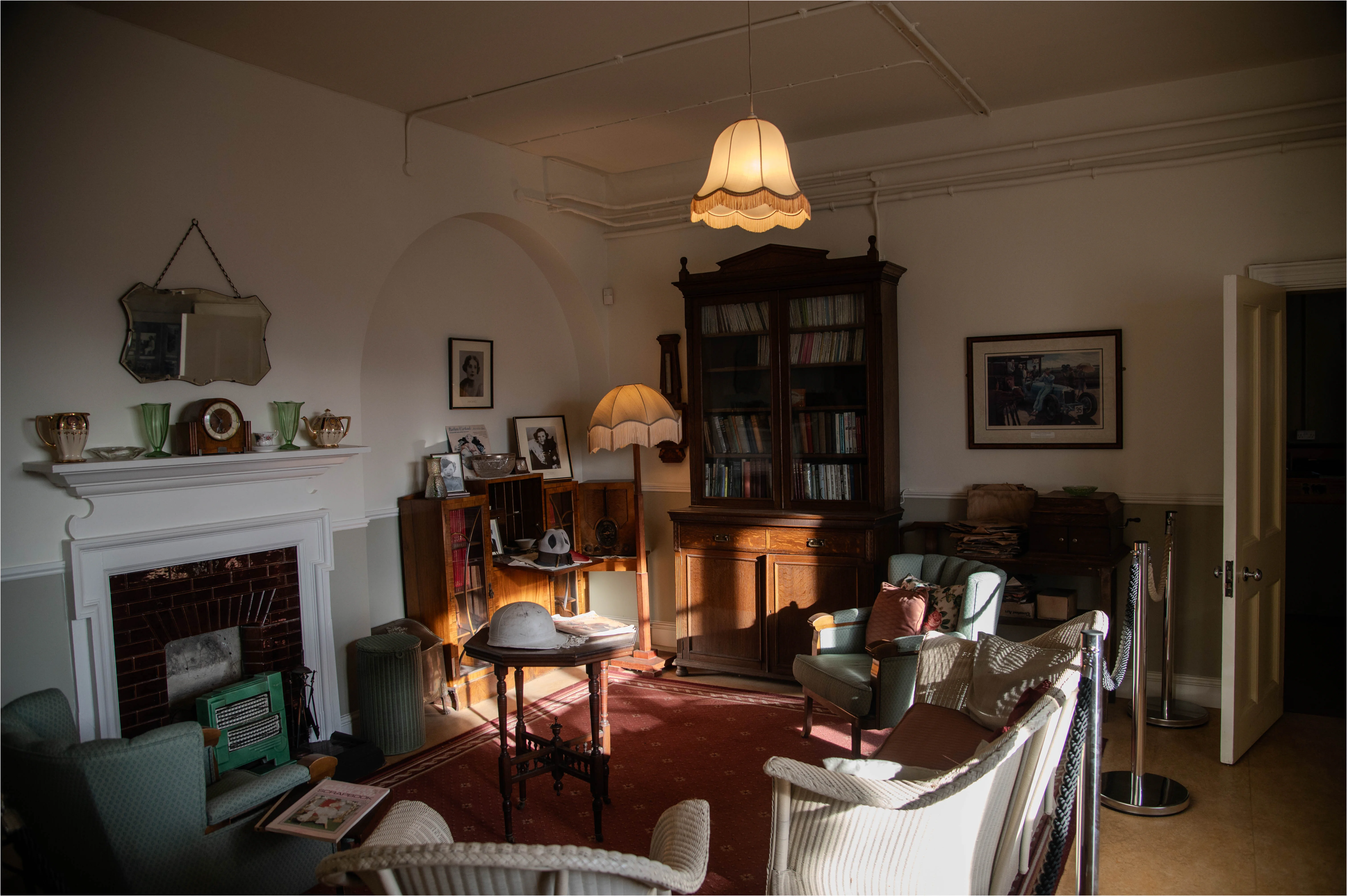 A dimly lit living room furnished in a vintage style, showcasing a fireplace, a large wooden bookcase, armchairs, and a coffee table with a hat on it.
