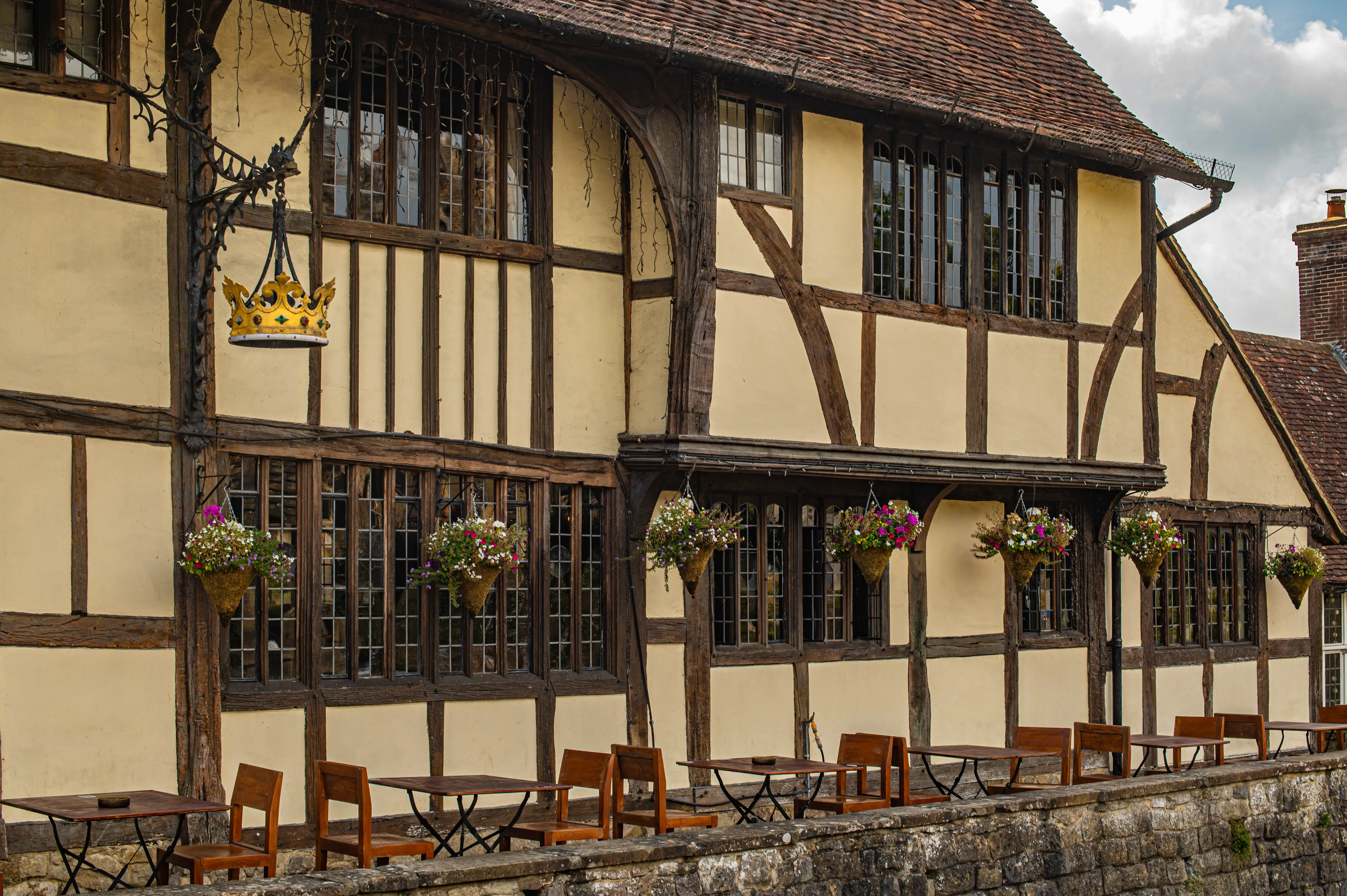 Timber-framed English building with golden crown sign, hanging flower baskets and outdoor seating.