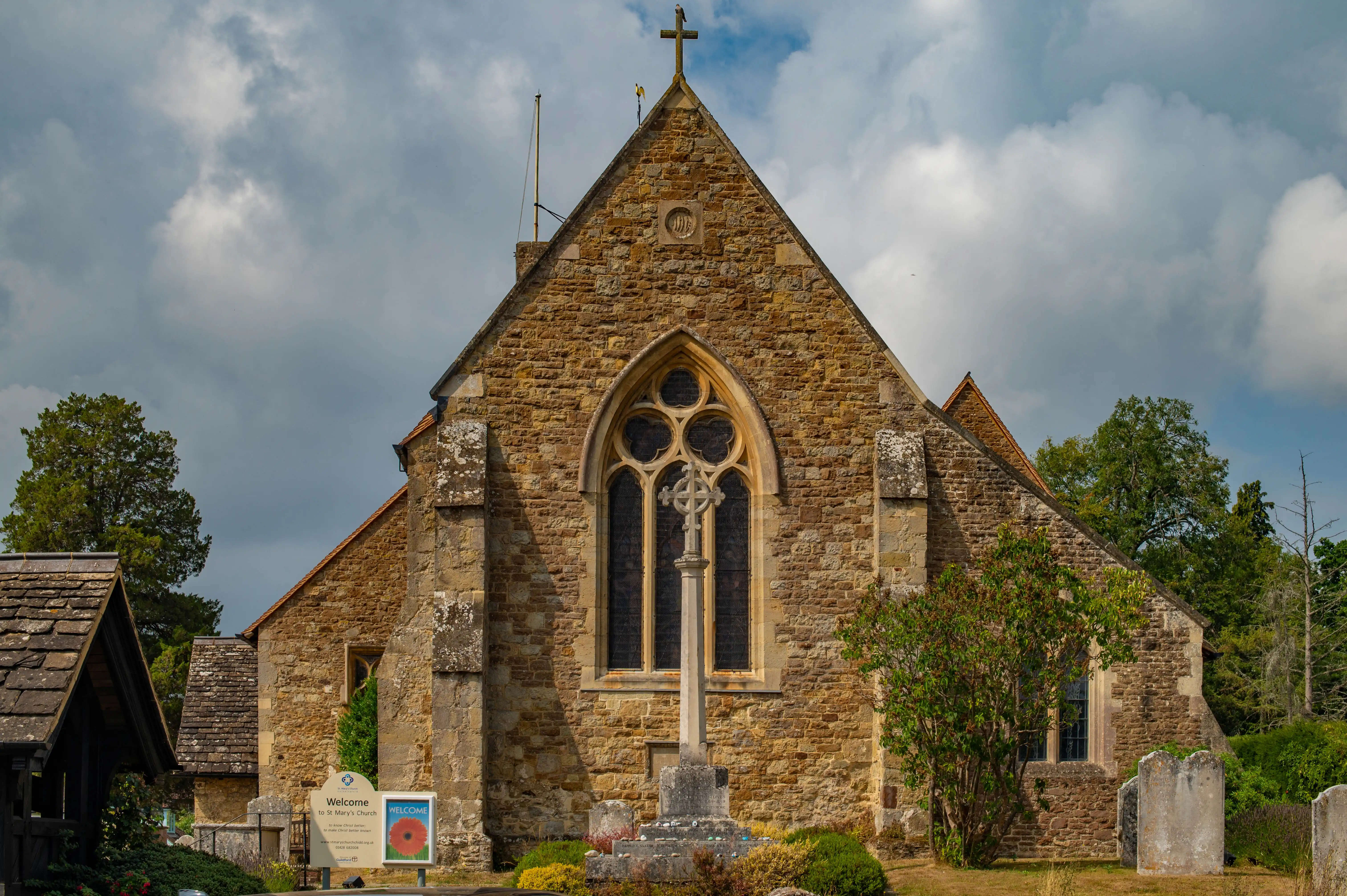 Stone church with arched stained-glass window, tall cross monument and gravestones in a churchyard.