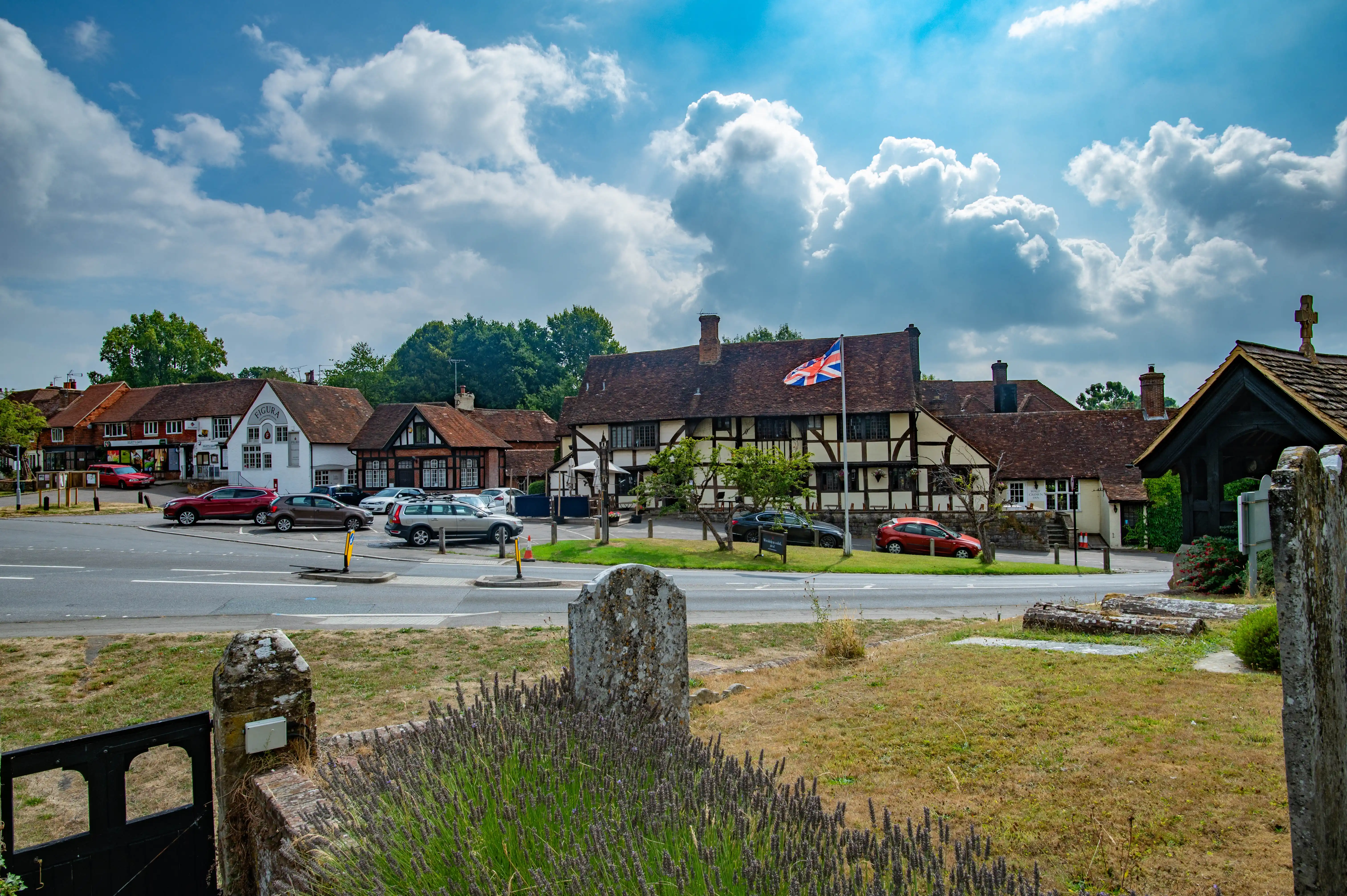 A village scene in England, featuring Tudor-style buildings, a Union Jack flag, a road with parked cars, and green trees under a cloudy sky.