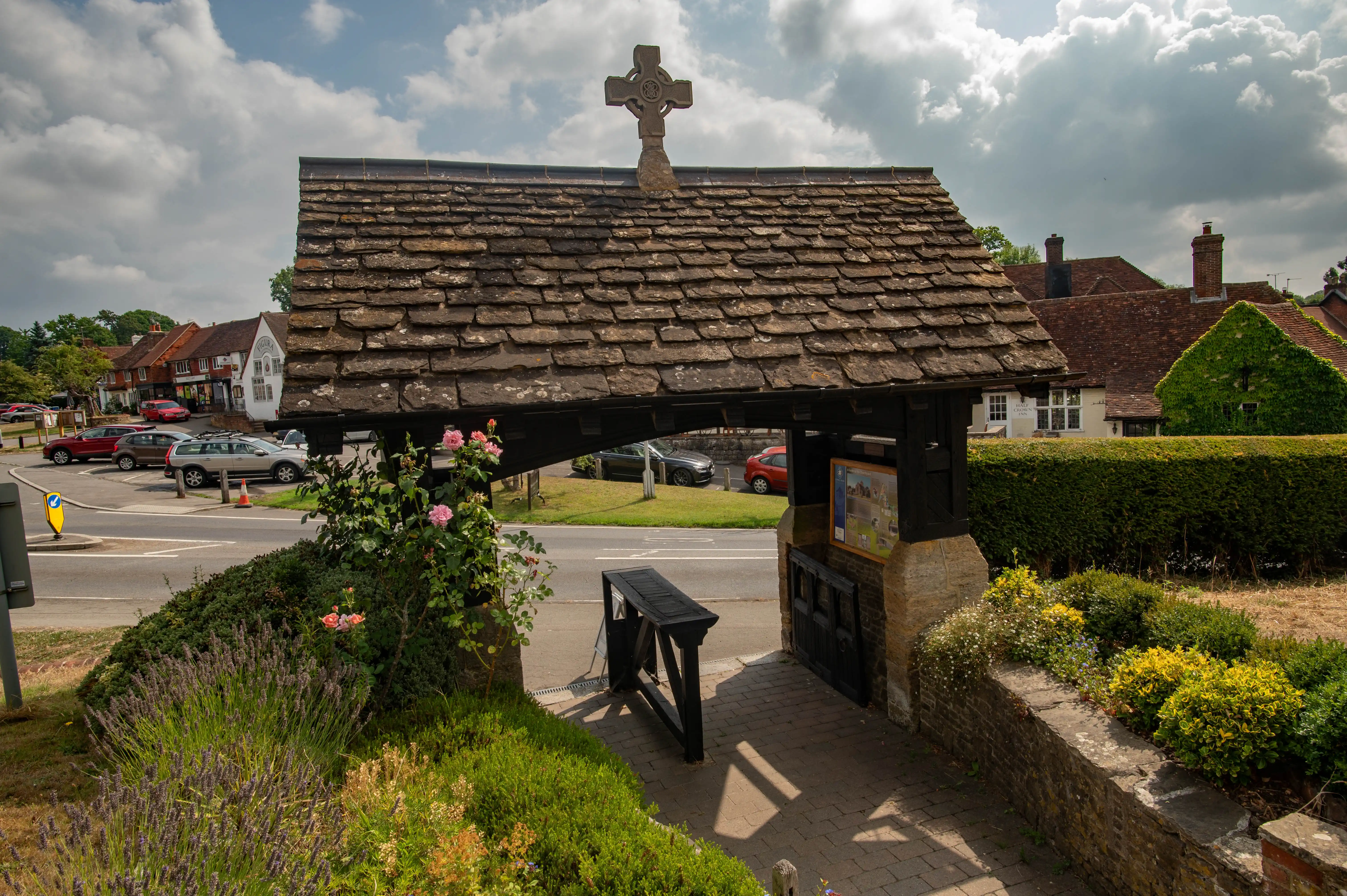 Lychgate with shingled roof and Celtic cross, surrounded by roses, with village buildings across the road.
