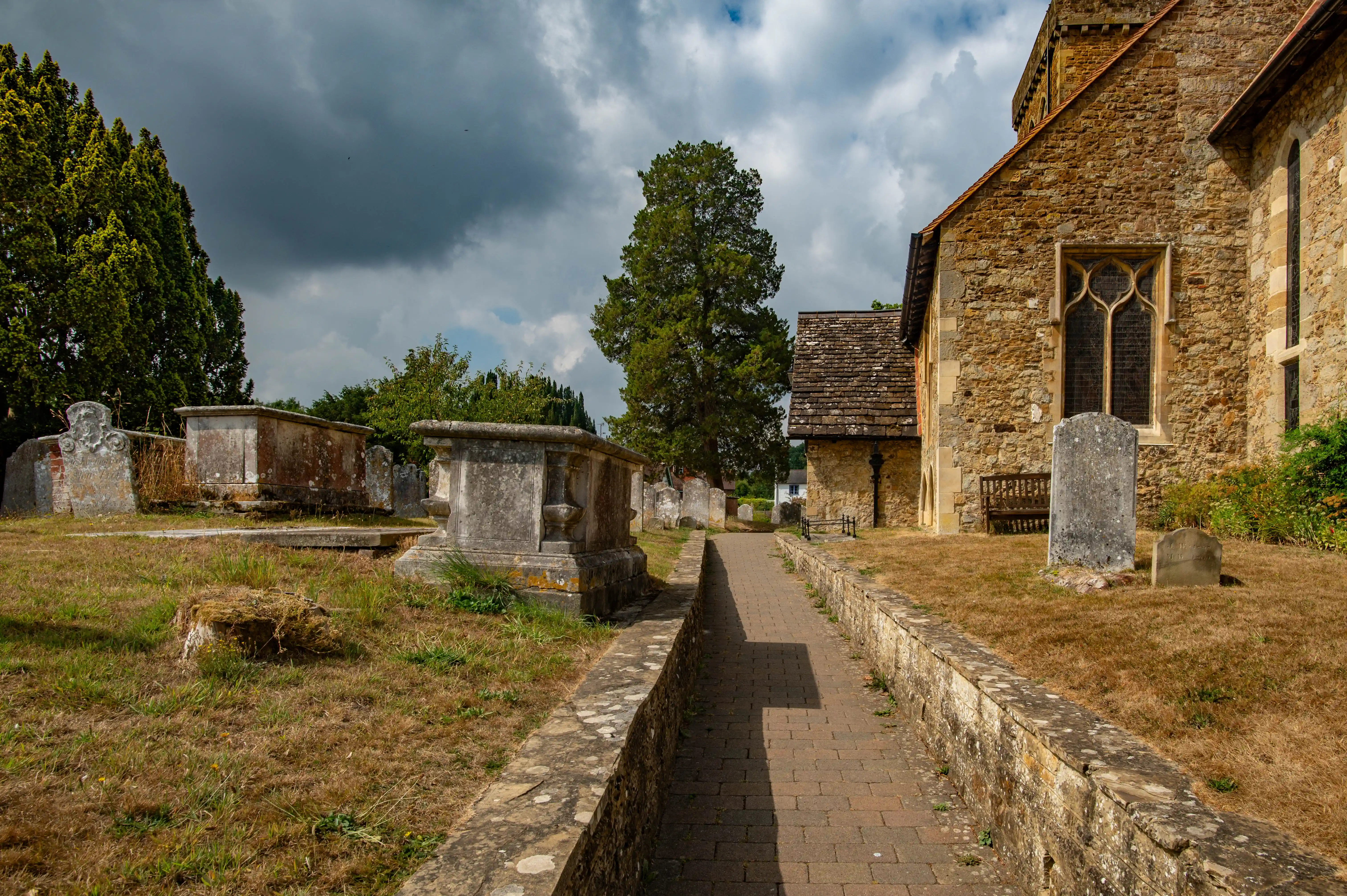 Stone church with arched windows, raised burial monuments and dry grass in a historic English churchyard.