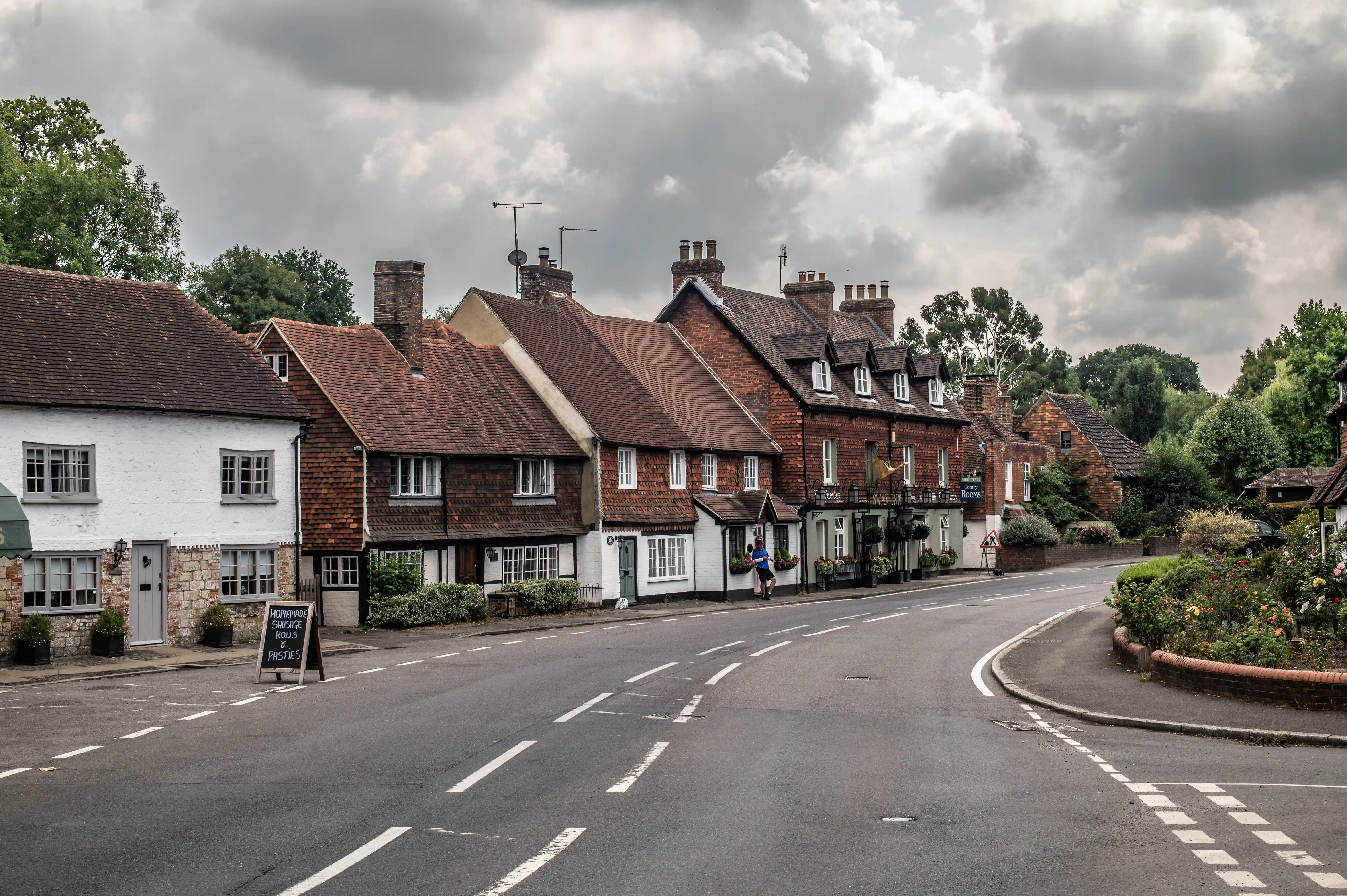 Curving English village street with timber-framed houses, garden flowers and a person walking under an overcast sky.