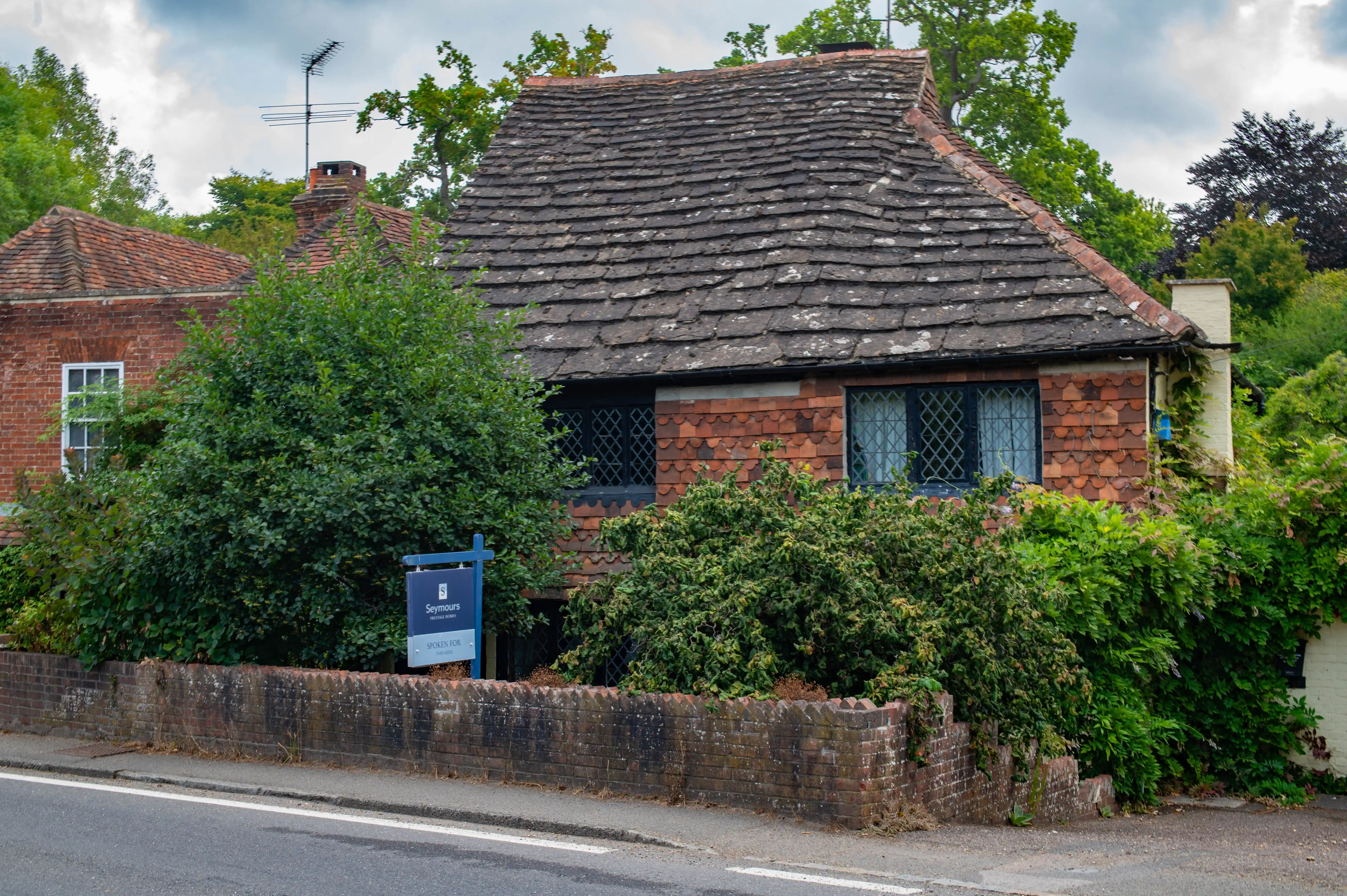 Small brick cottage with steep tiled roof, leaded windows, overgrown greenery and a “For Sale” sign.