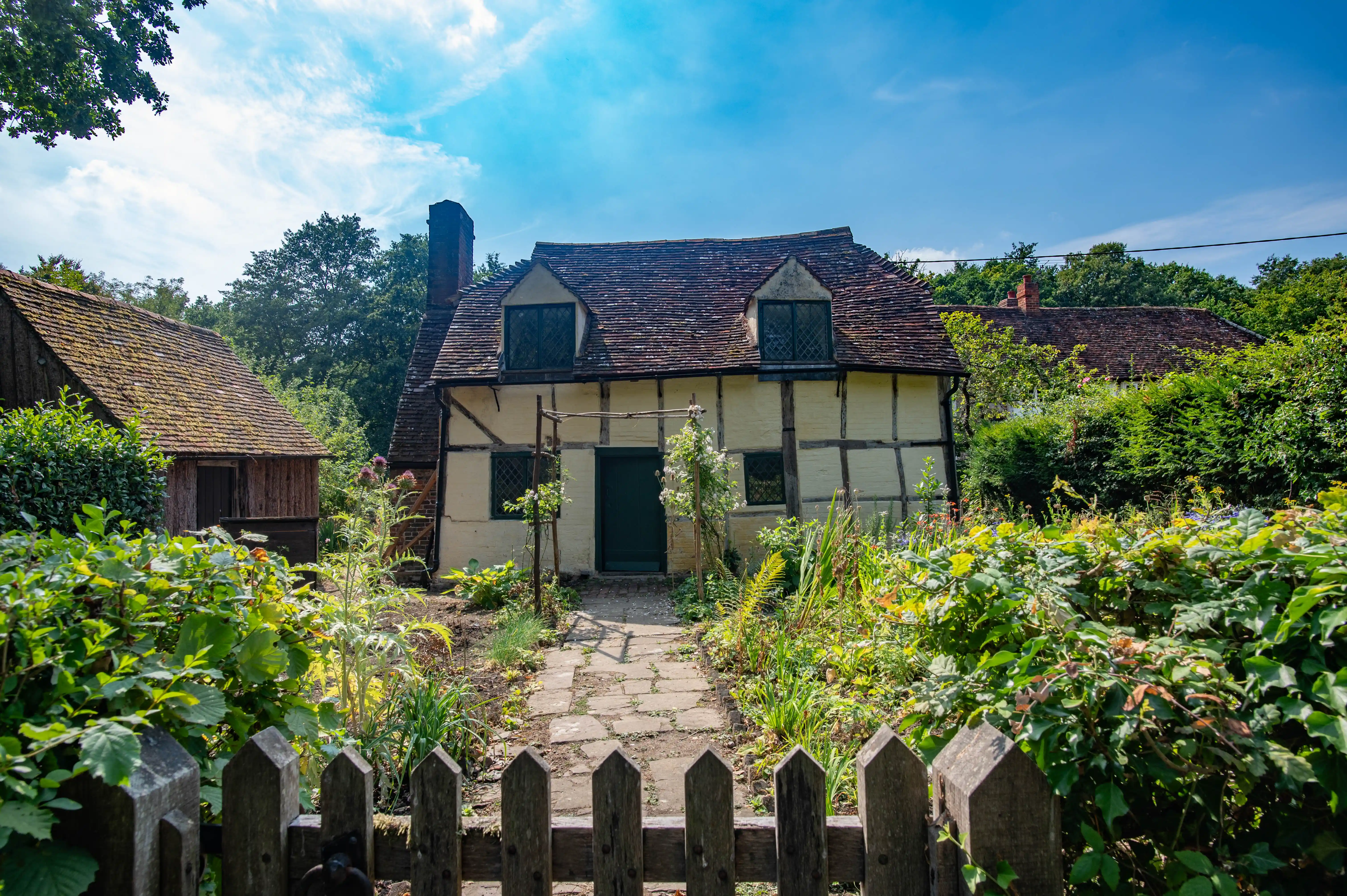 A half-timbered cottage with a thatched roof, a stone path leading to the front door, and a wooden fence in the foreground, set against a backdrop of trees and a blue sky.
