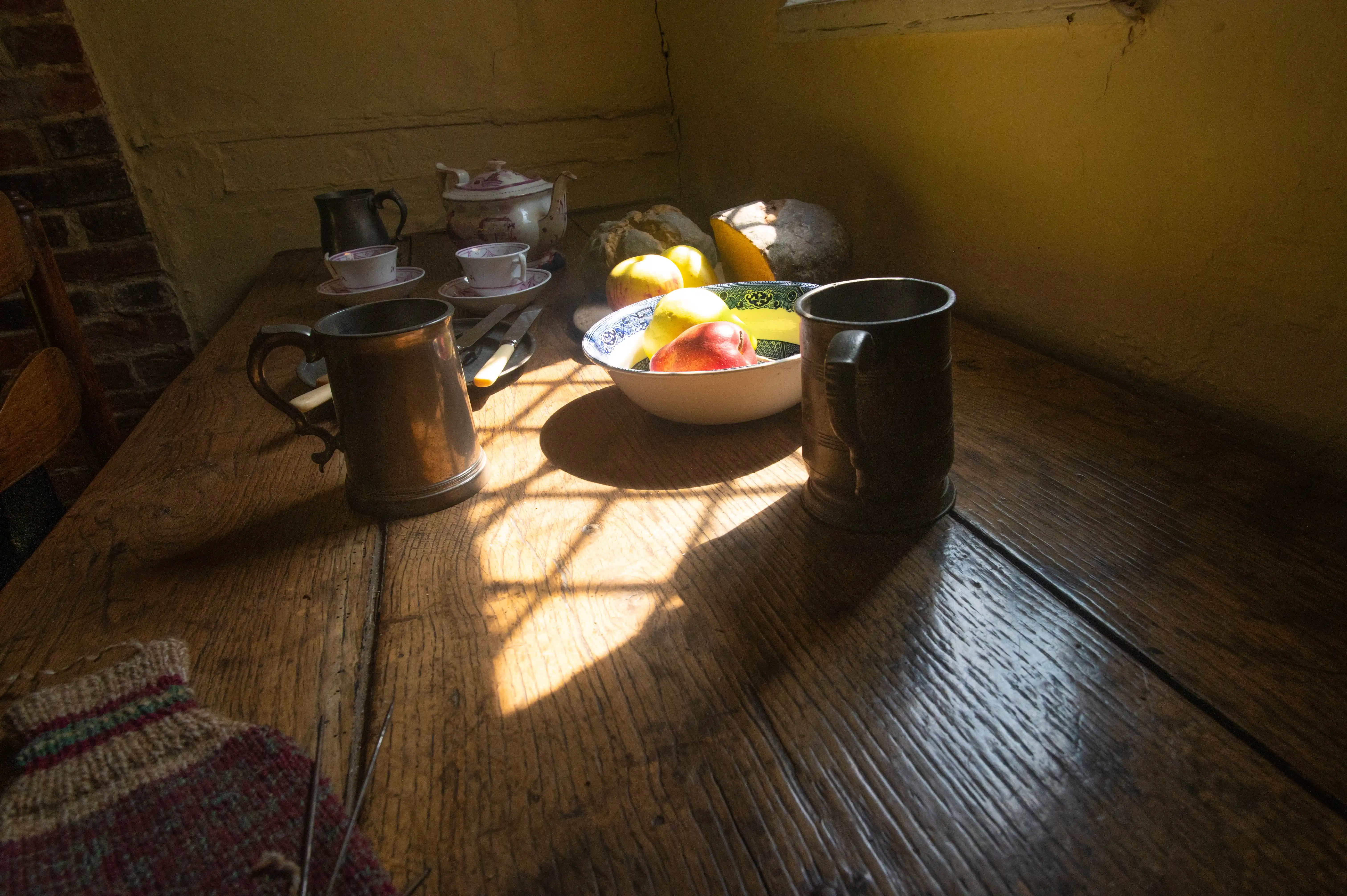 A low-angle view of a weathered wooden table set with a bowl of apples and pears, two metal tankards, a teapot, and cups, with dramatic sunlight casting a grid pattern shadow.