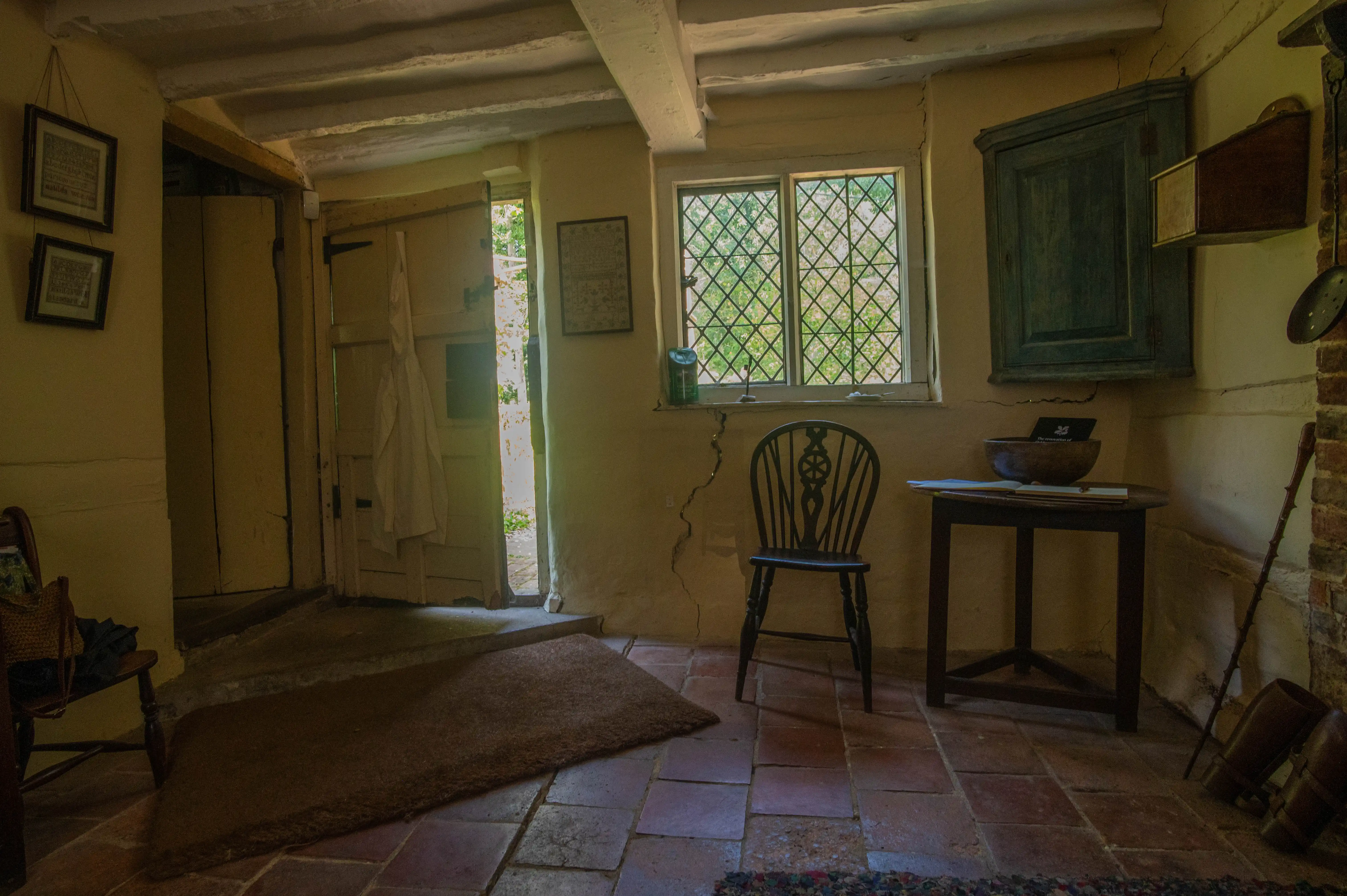 A dimly lit interior of a rustic cottage with a wooden door ajar, revealing bright sunlight. A diamond-paned lattice window is visible on the right wall, with a dark wooden chair in front of it. A small wooden table with a bowl and book stands next to the chair. Terracotta floor tiles and exposed wooden beams on the ceiling complete the scene.