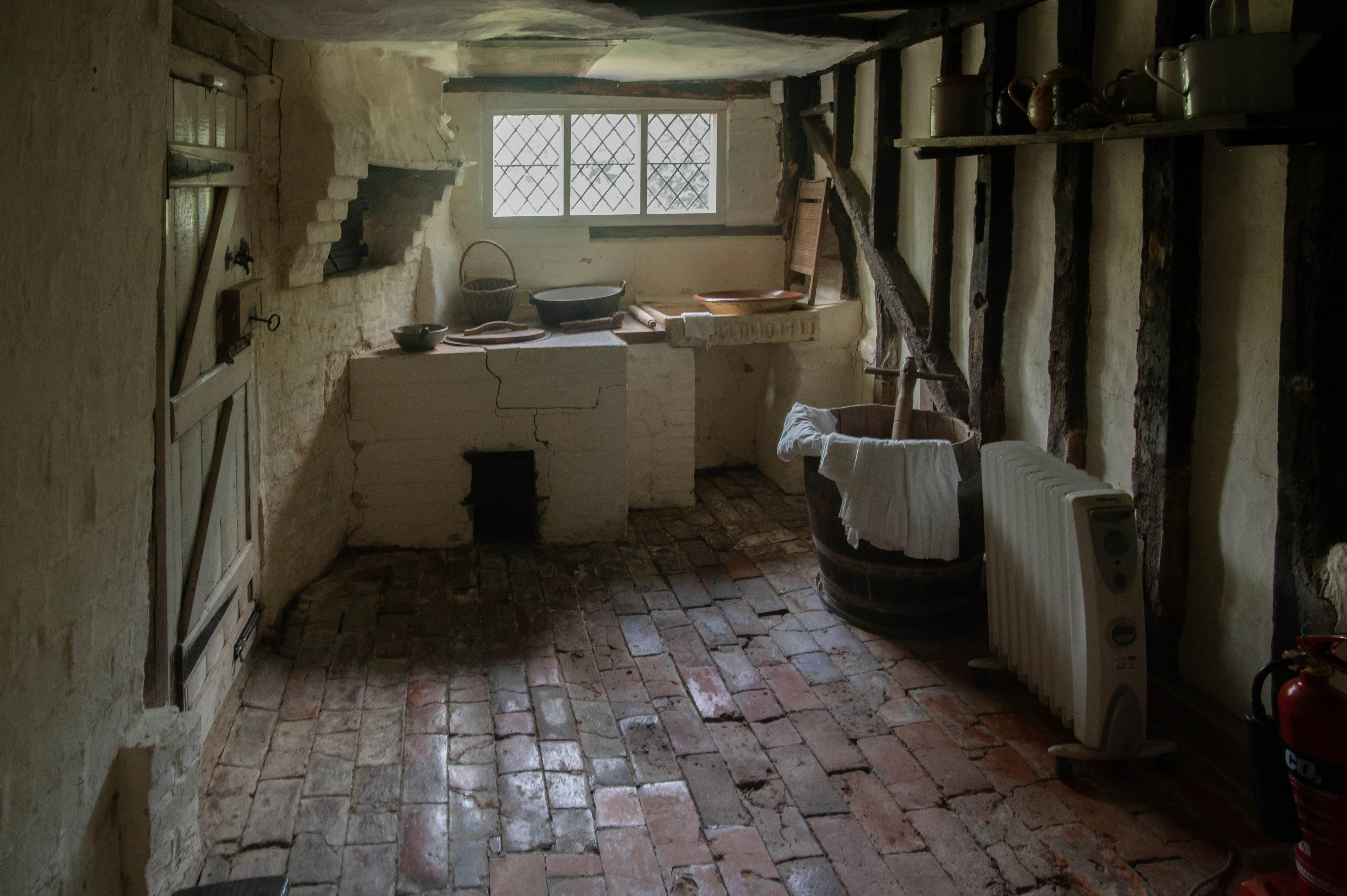 Interior view of a rustic historical kitchen with whitewashed walls, exposed dark timber beams, a traditional brick fireplace, and a flagstone floor. A wooden door is on the left, and a window with diamond panes is above the hearth. A wooden wash barrel with white cloth, an electric heater, and a red fire extinguisher are visible on the right.