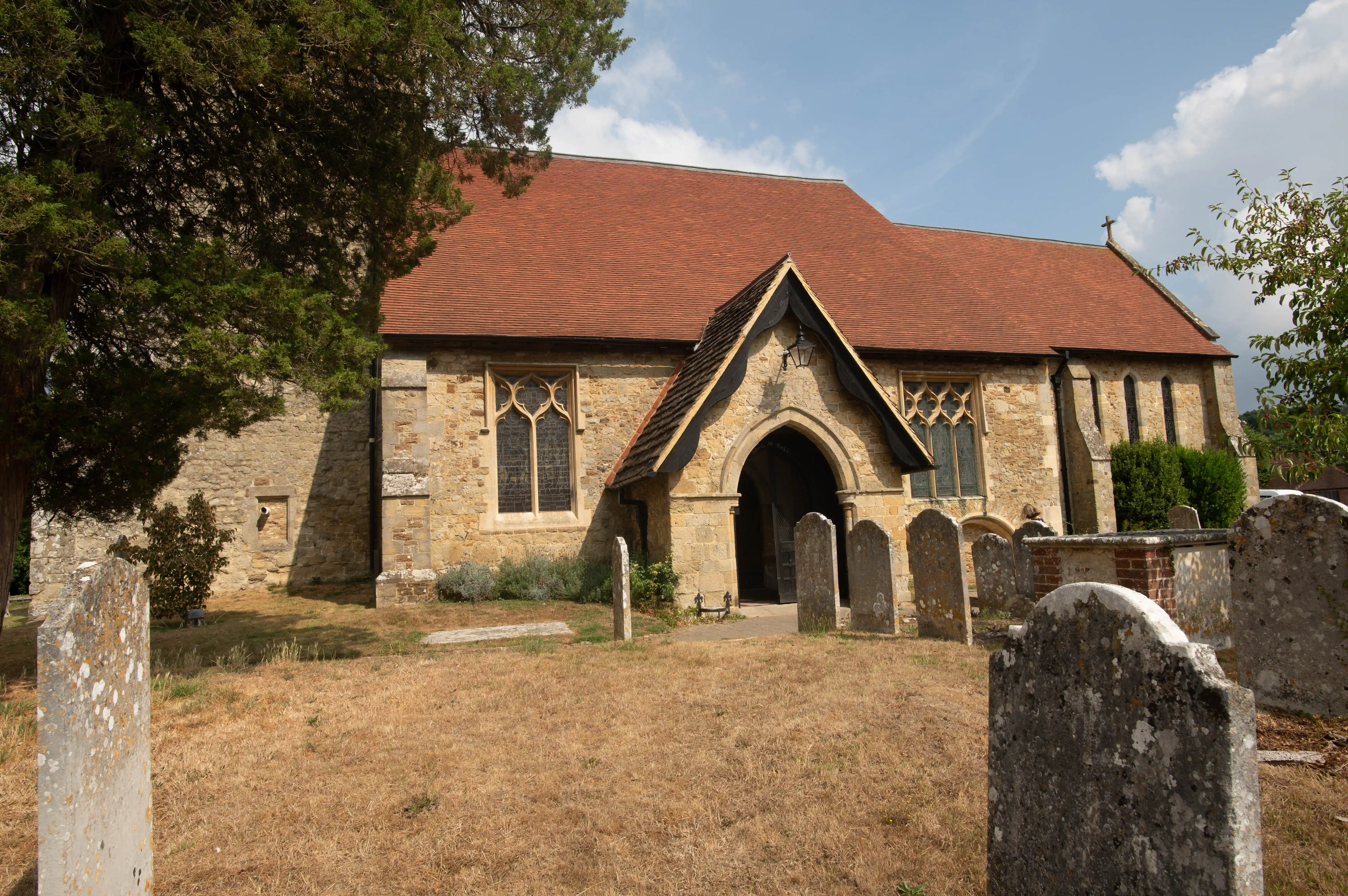 Stone church with red-tiled roof, Gothic windows, arched entrance and weathered headstones in a churchyard.