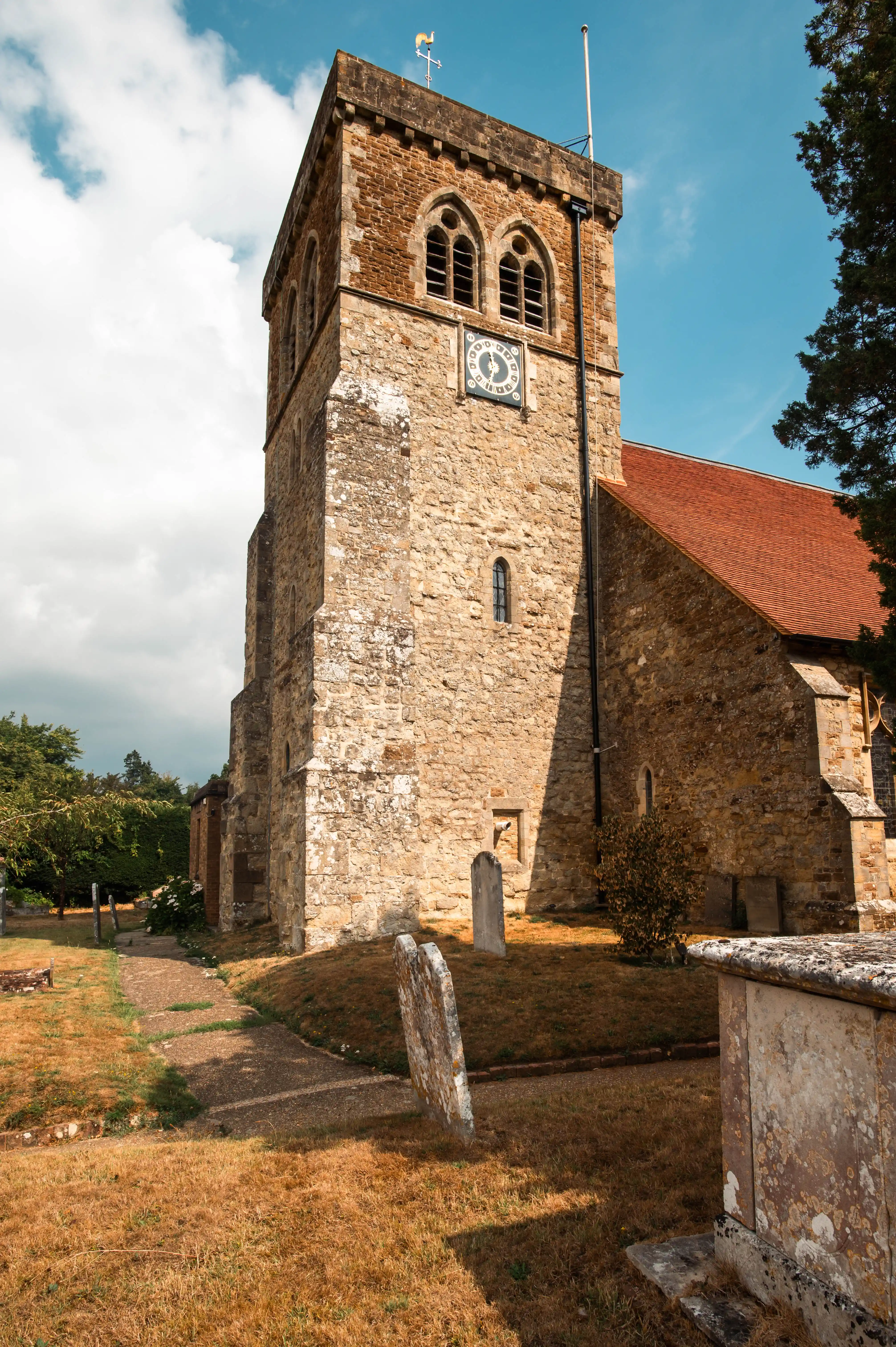Stone church with square clock tower, arched windows and gravestones in a historic English churchyard.