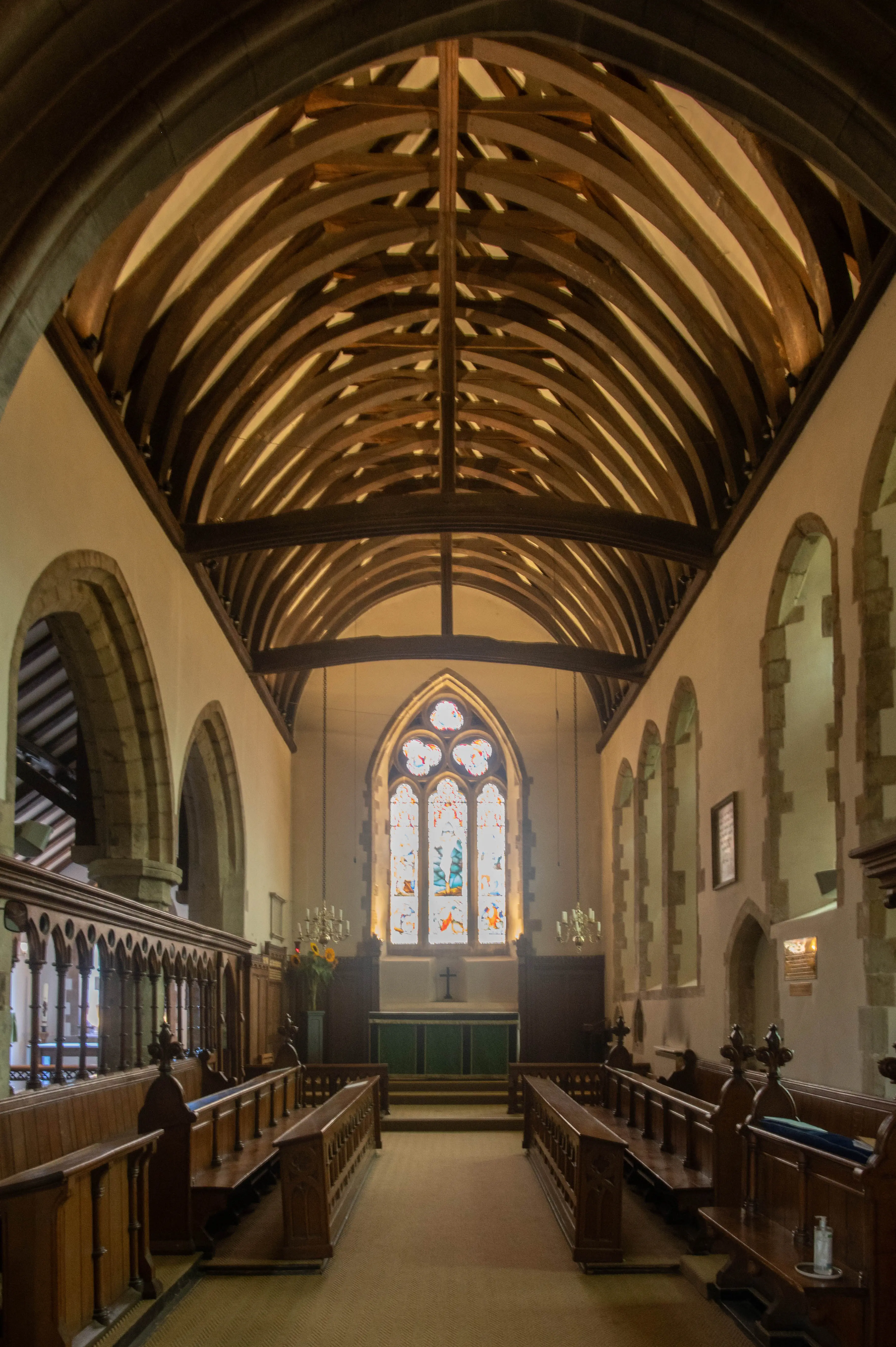Interior of a church with wooden pews lining the nave, a high vaulted wooden ceiling, and a large stained glass window above the altar.