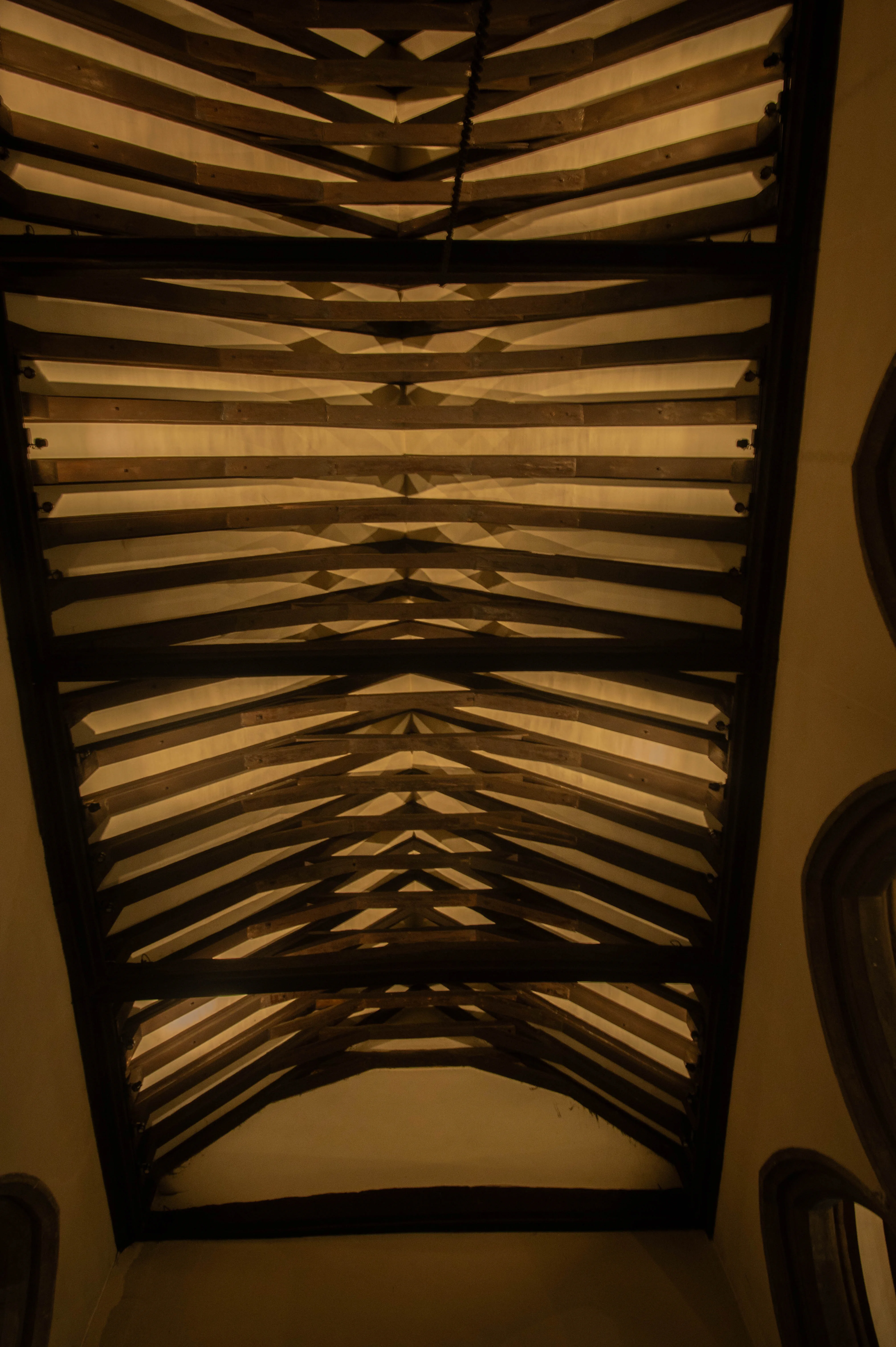 A close-up, low-angle view of a dark wooden beam ceiling. The beams form repeating 'V' shapes that converge towards the centre, with warm yellow light illuminating the spaces between them and creating stark shadows. The edge of a pale wall is visible on the right.
