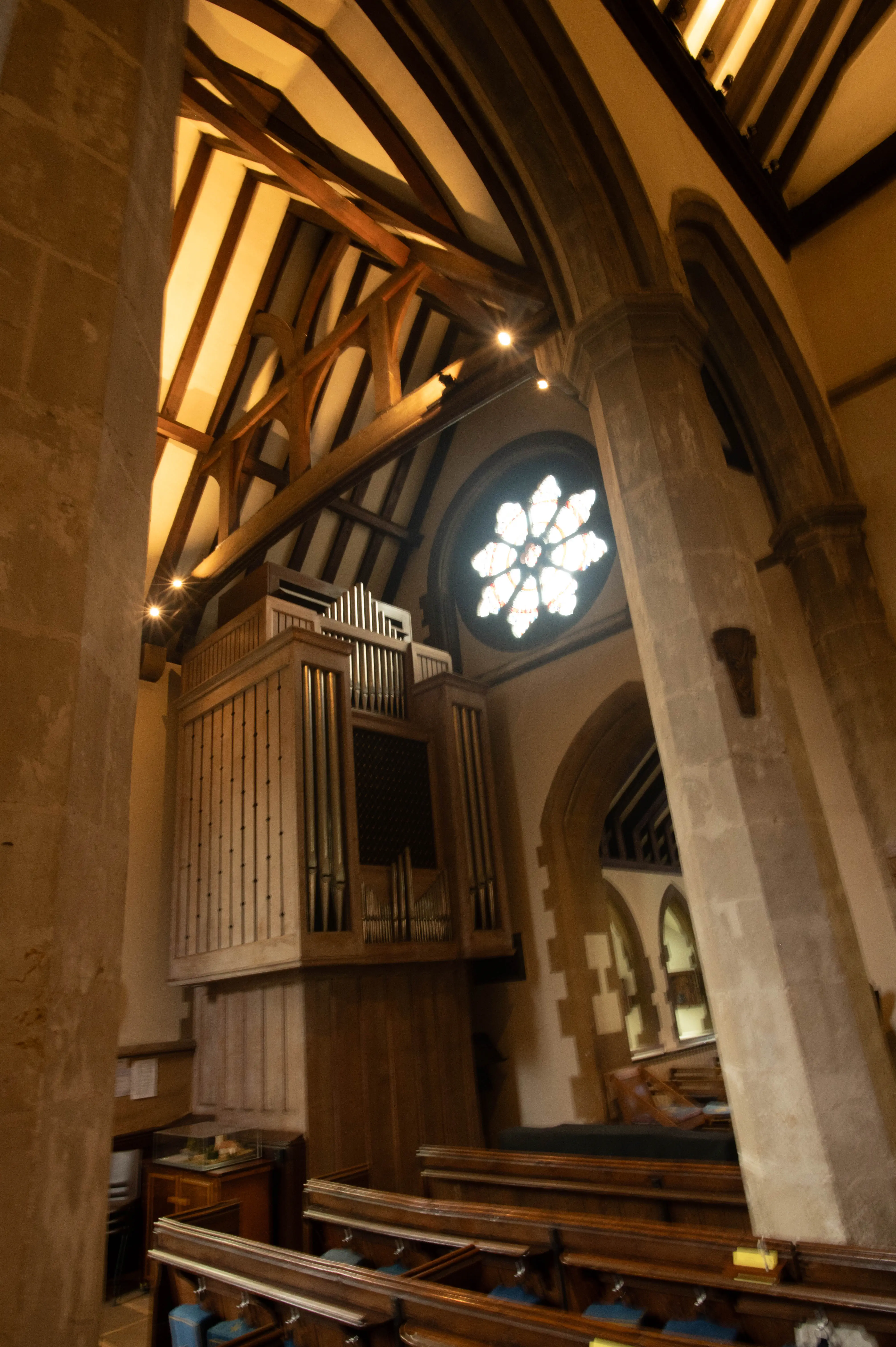Interior view of a church with a large wooden pipe organ on the left, a circular rose window at the top, and rows of wooden pews in the foreground. The ceiling features exposed wooden beams.