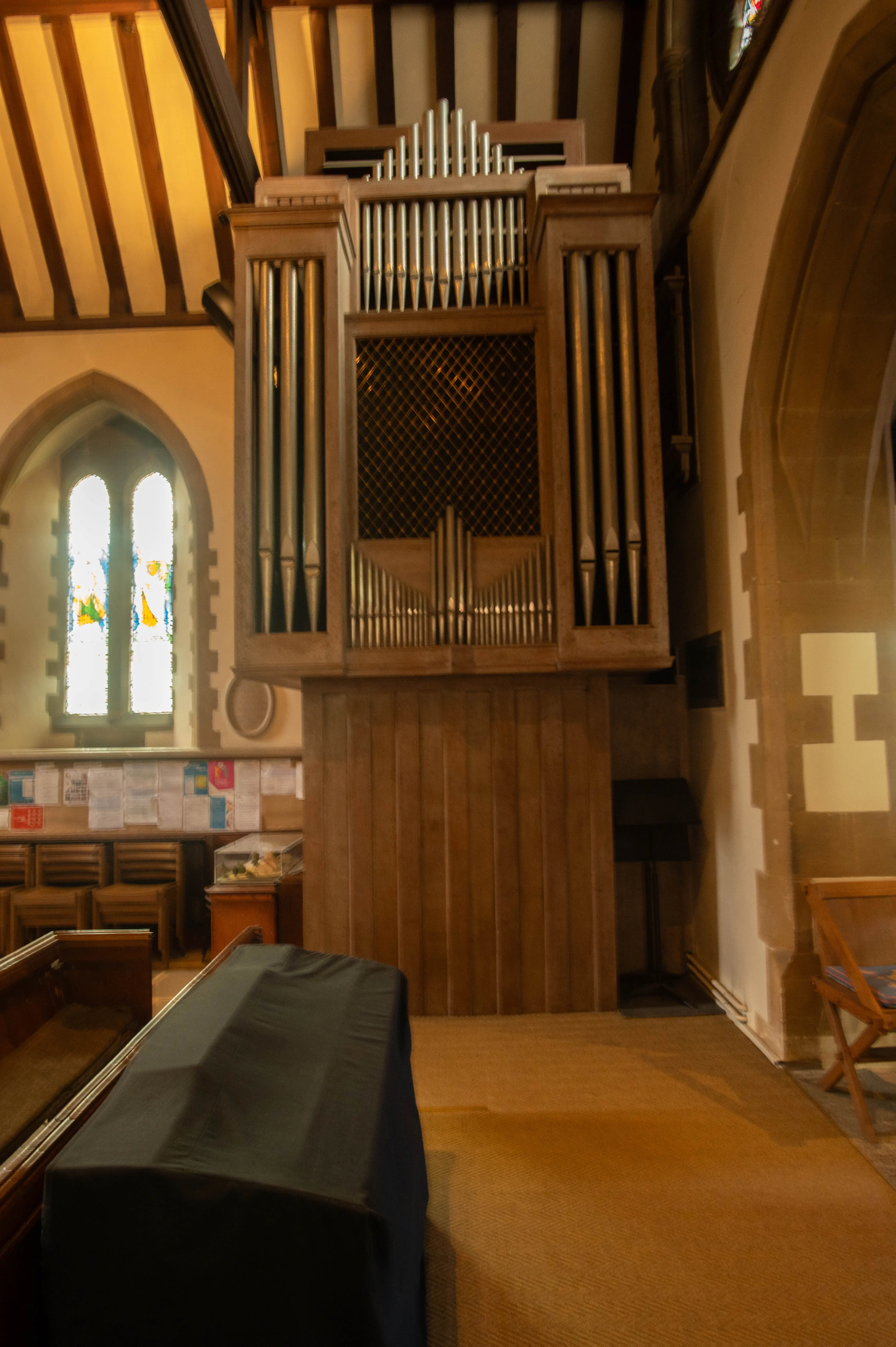 A large pipe organ with many metal pipes is situated in a church. The organ has a wooden casing and stands against a wall. To the left is a tall arched window with light streaming through. In the foreground, pews and a large object covered in a black cloth are visible.