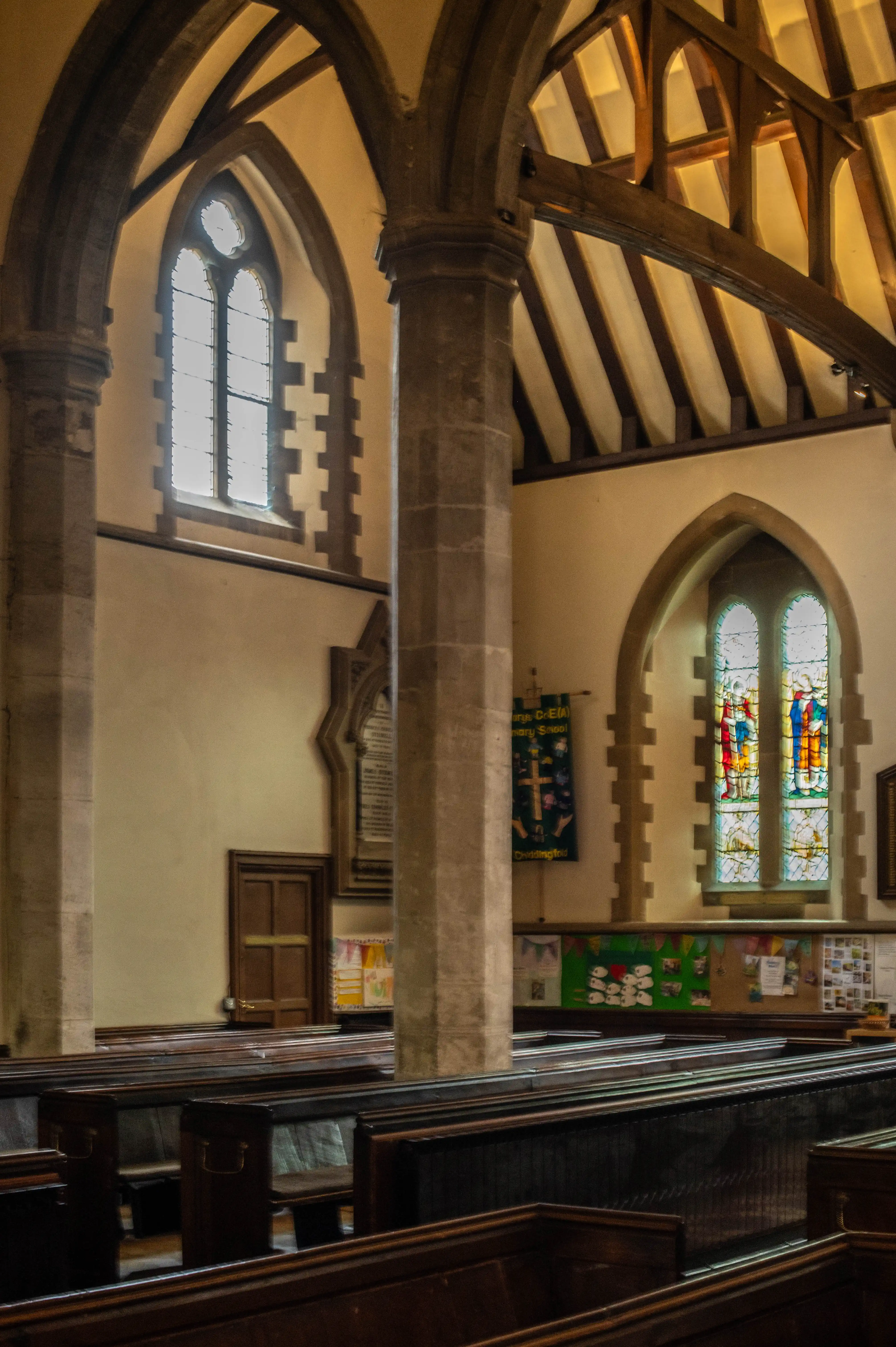 Church interior with stone columns, stained-glass windows, wooden beams and pews under warm lighting.
