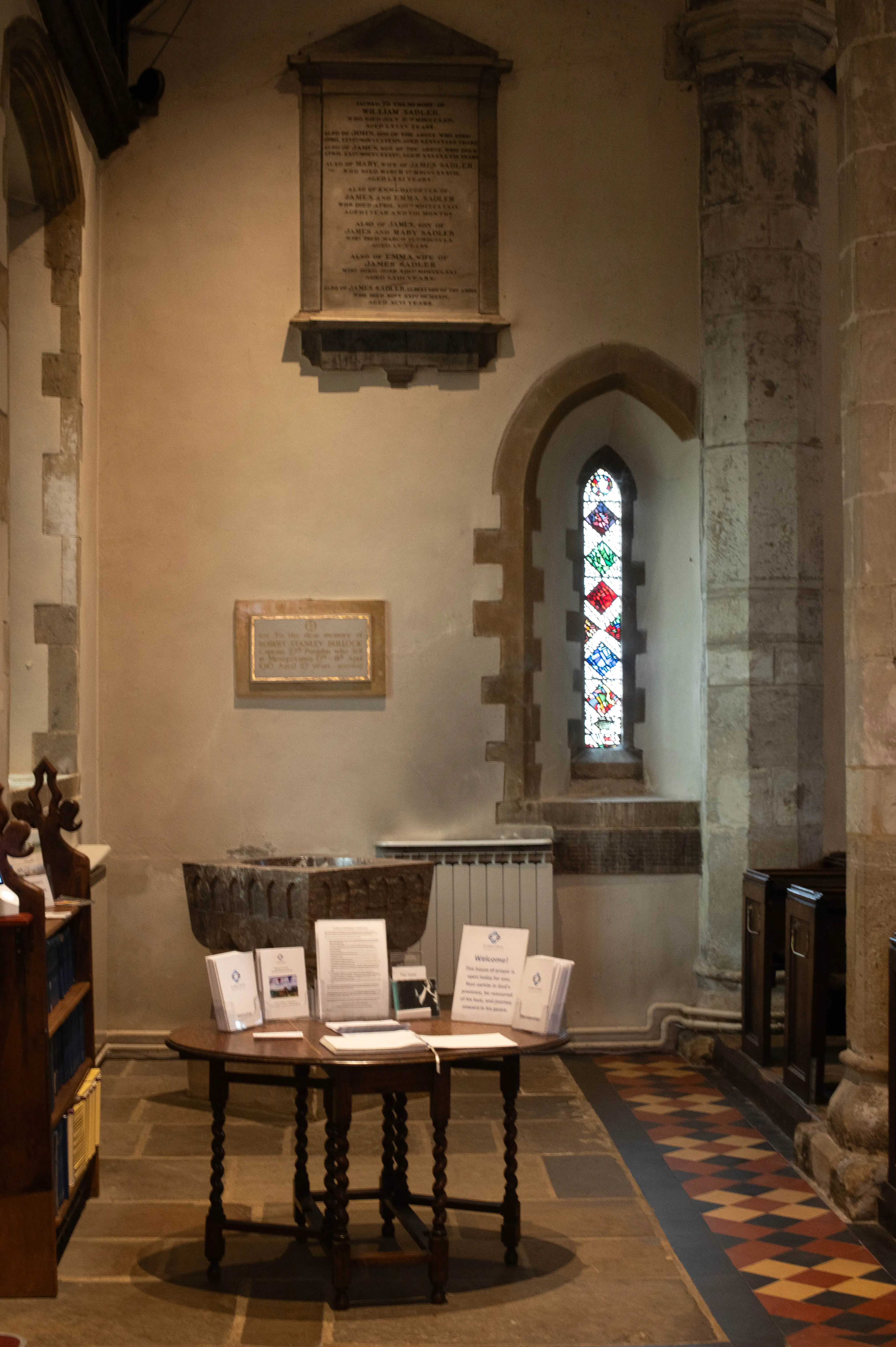 Church interior with geometric stained-glass window, memorial plaques, stone floor and wooden table.