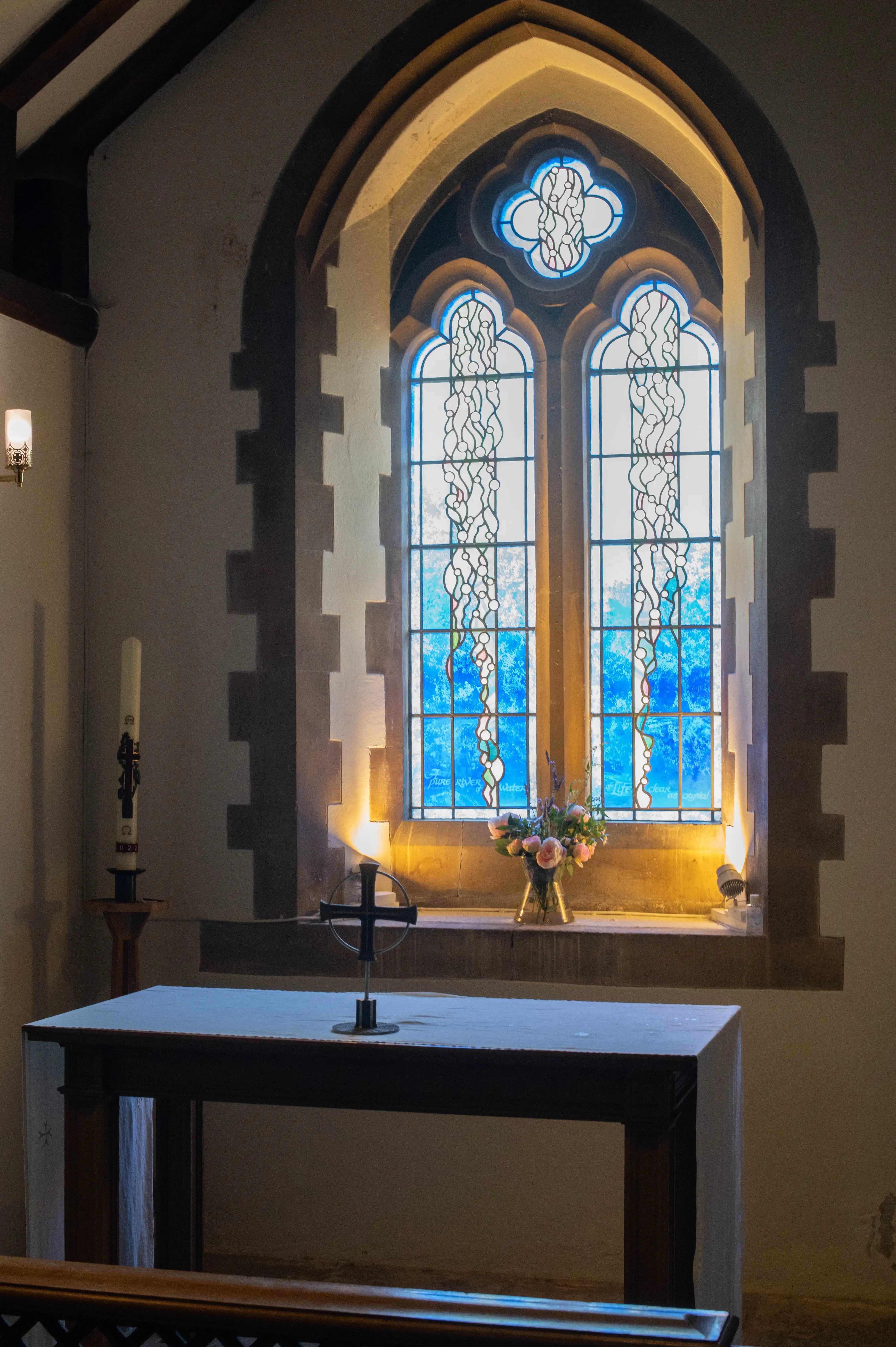 Chapel interior with blue stained-glass window, stone ledge with flowers, altar cloth, cross ornament and tall candle.