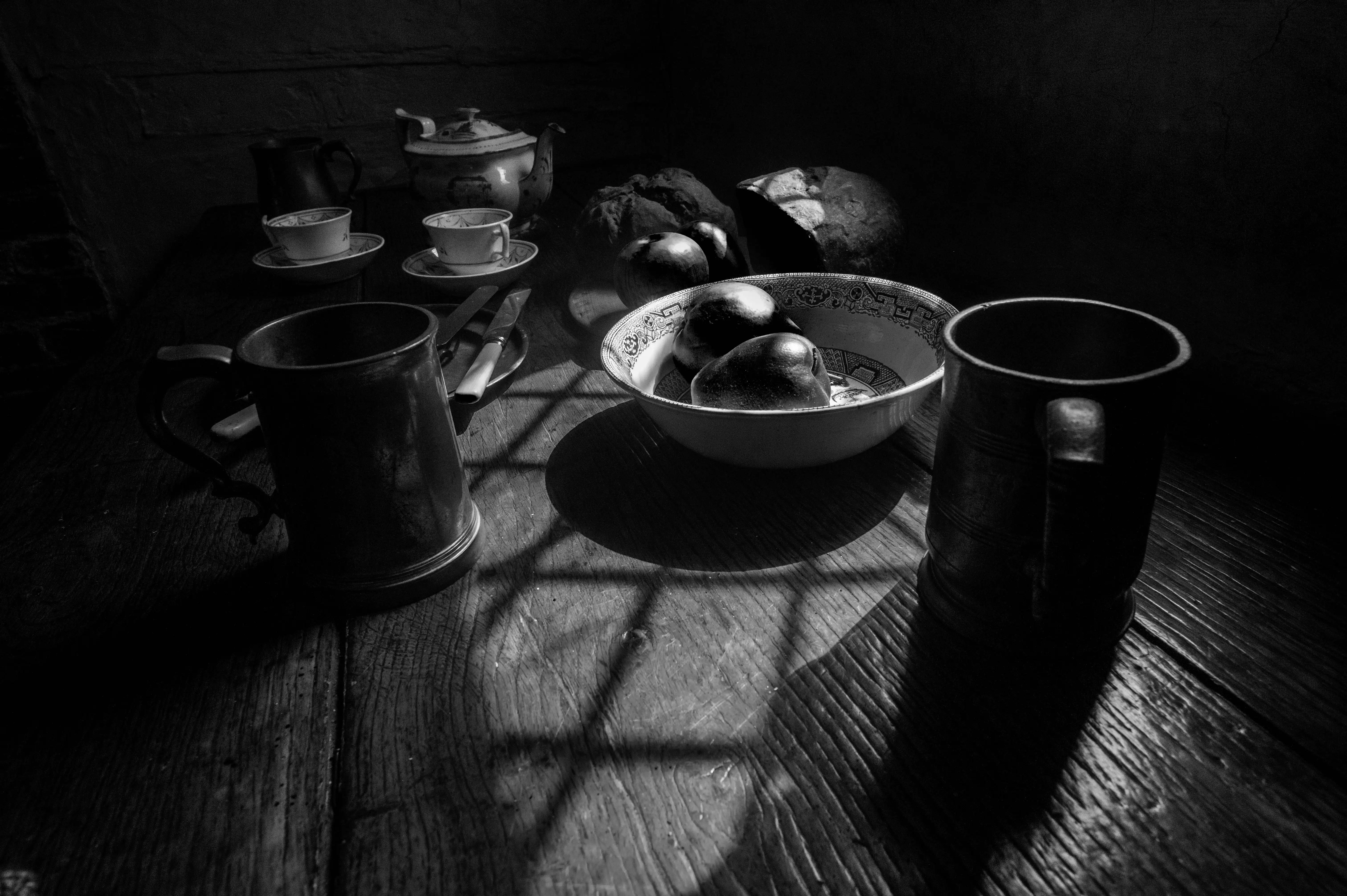 A black and white still life photograph showing a wooden table with two pewter tankards, teacups, saucers, a bowl of fruit including pears and apples, and bread. Light streams through a window, casting grid-like shadows across the scene.
