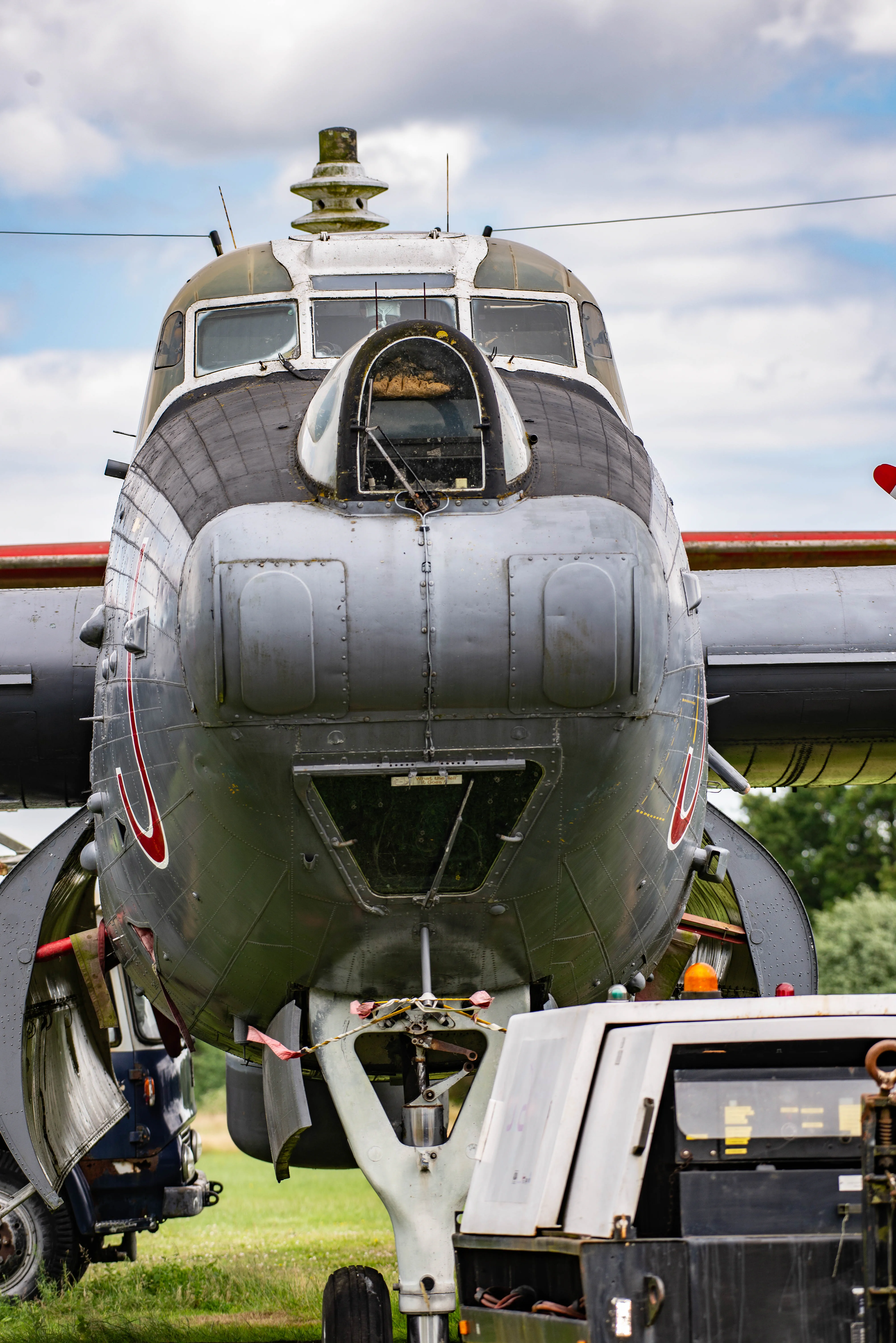 A close-up, frontal view of a grey vintage aircraft, focusing on its cockpit windows, nose cone, and landing gear. Parts of the wings and a nearby vehicle are also visible, suggesting it might be on display or undergoing maintenance in an outdoor setting.