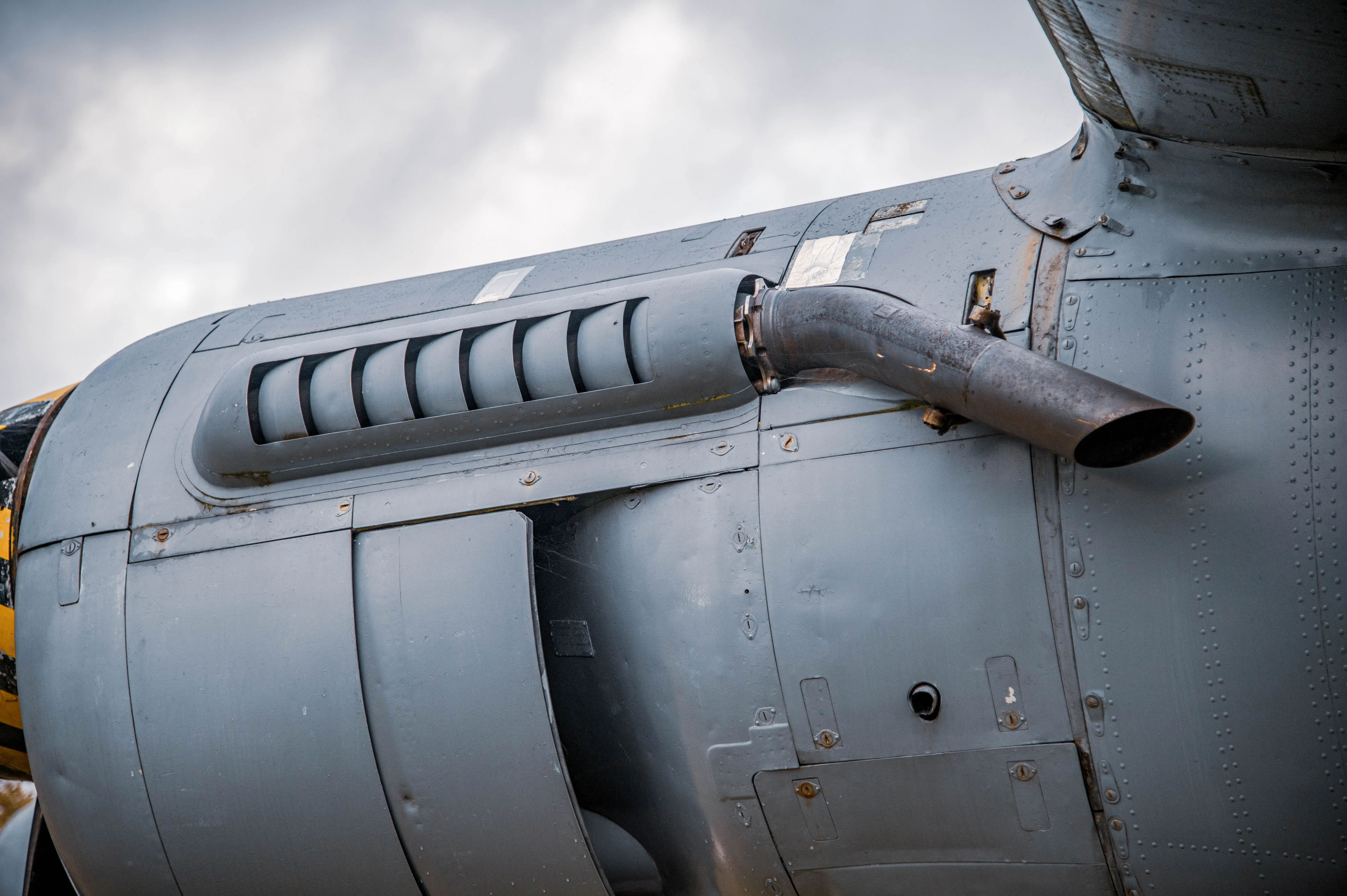 Close-up of the grey metal fuselage of an aircraft, showing a rusty exhaust pipe and a ventilation grille.