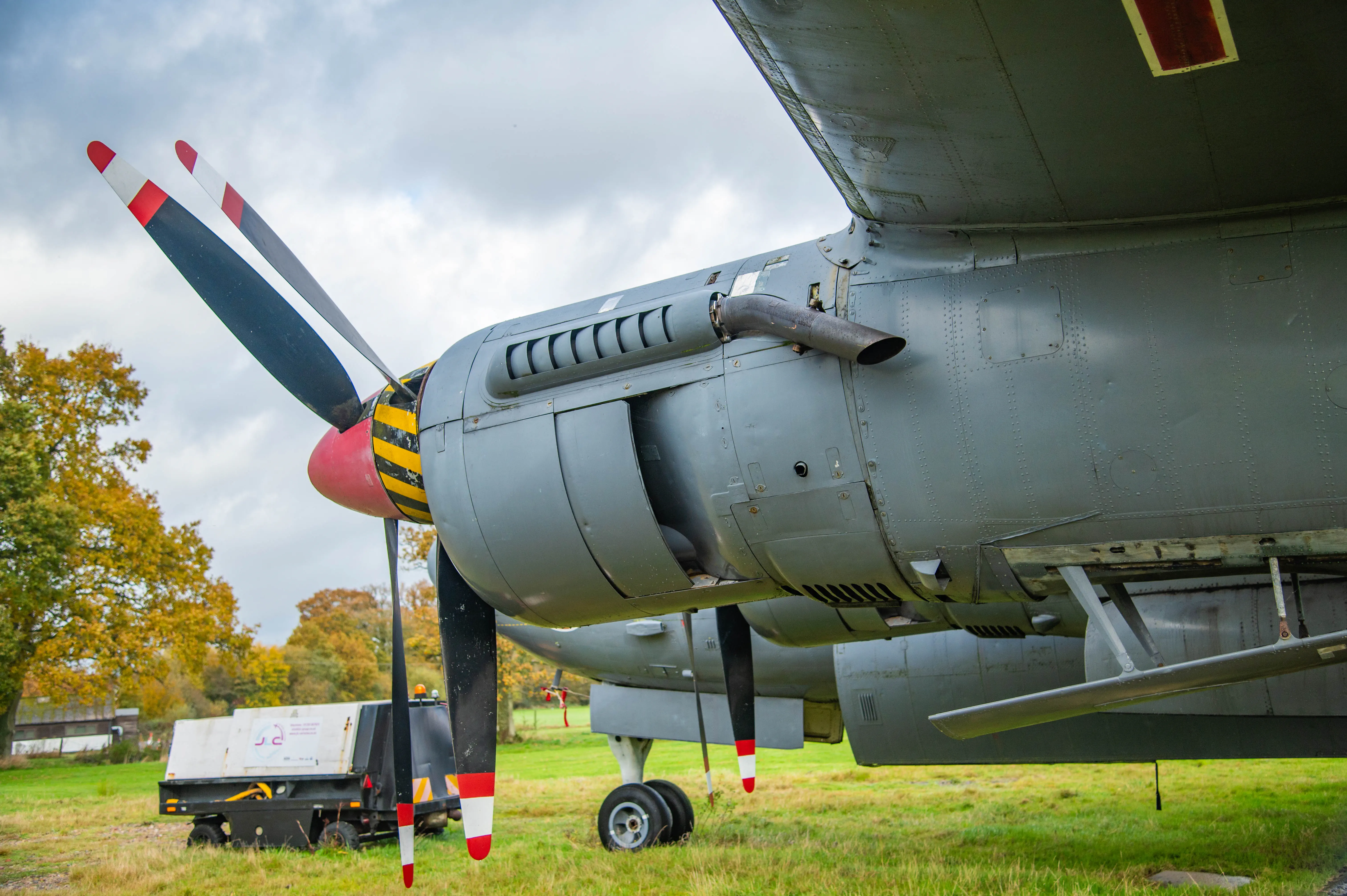 Close-up of a vintage aircraft engine nacelle with red spinner, striped propeller tips and exhaust outlet, parked on grass.