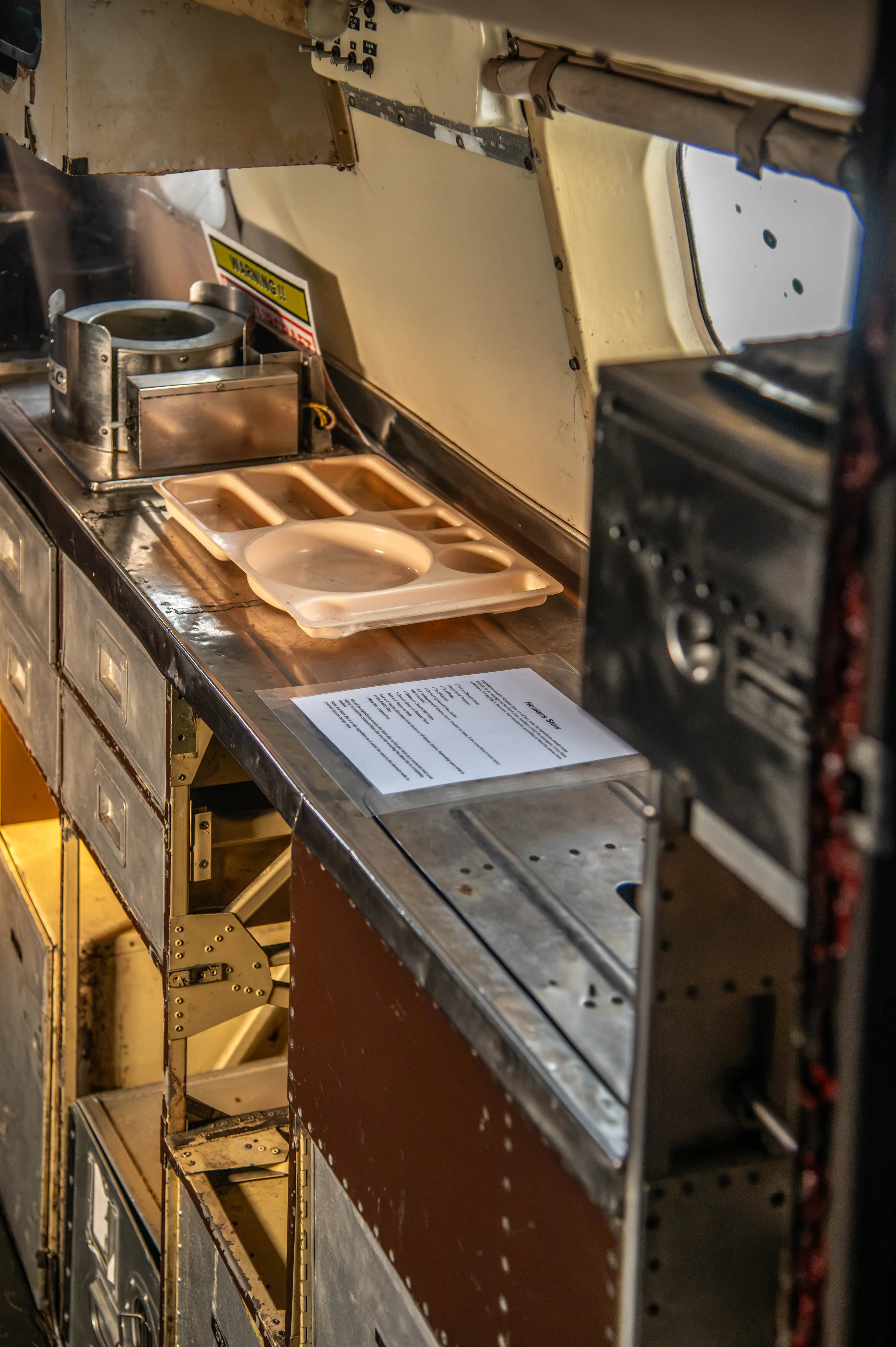 Close-up of a vintage aircraft galley counter with a beige meal tray, a metal food warmer, and a printed recipe titled 
