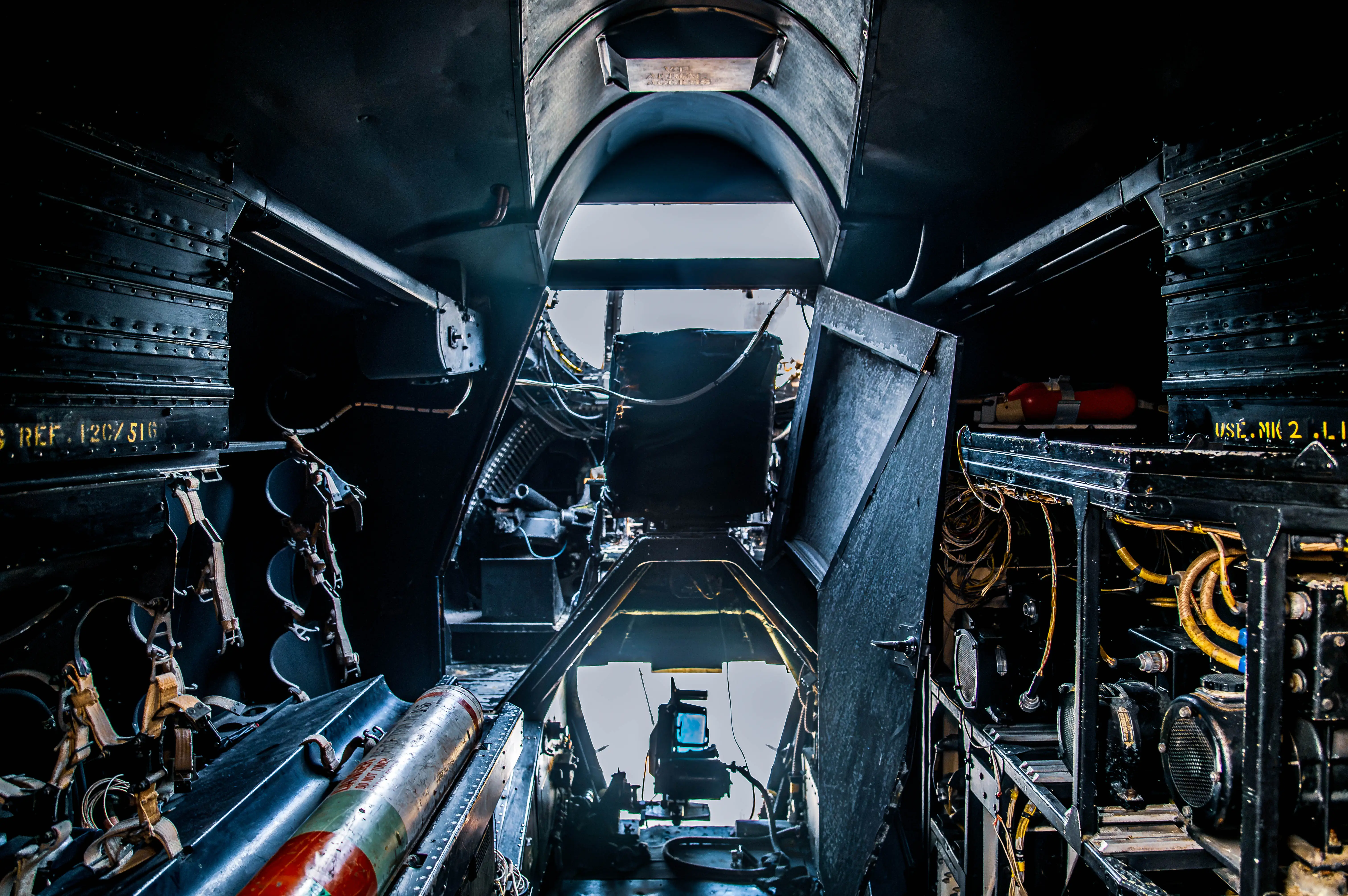 A dark, high-contrast view from inside the bomb bay of an old bomber aircraft, showing rows of bomb racks, intricate wiring, and mechanical components.