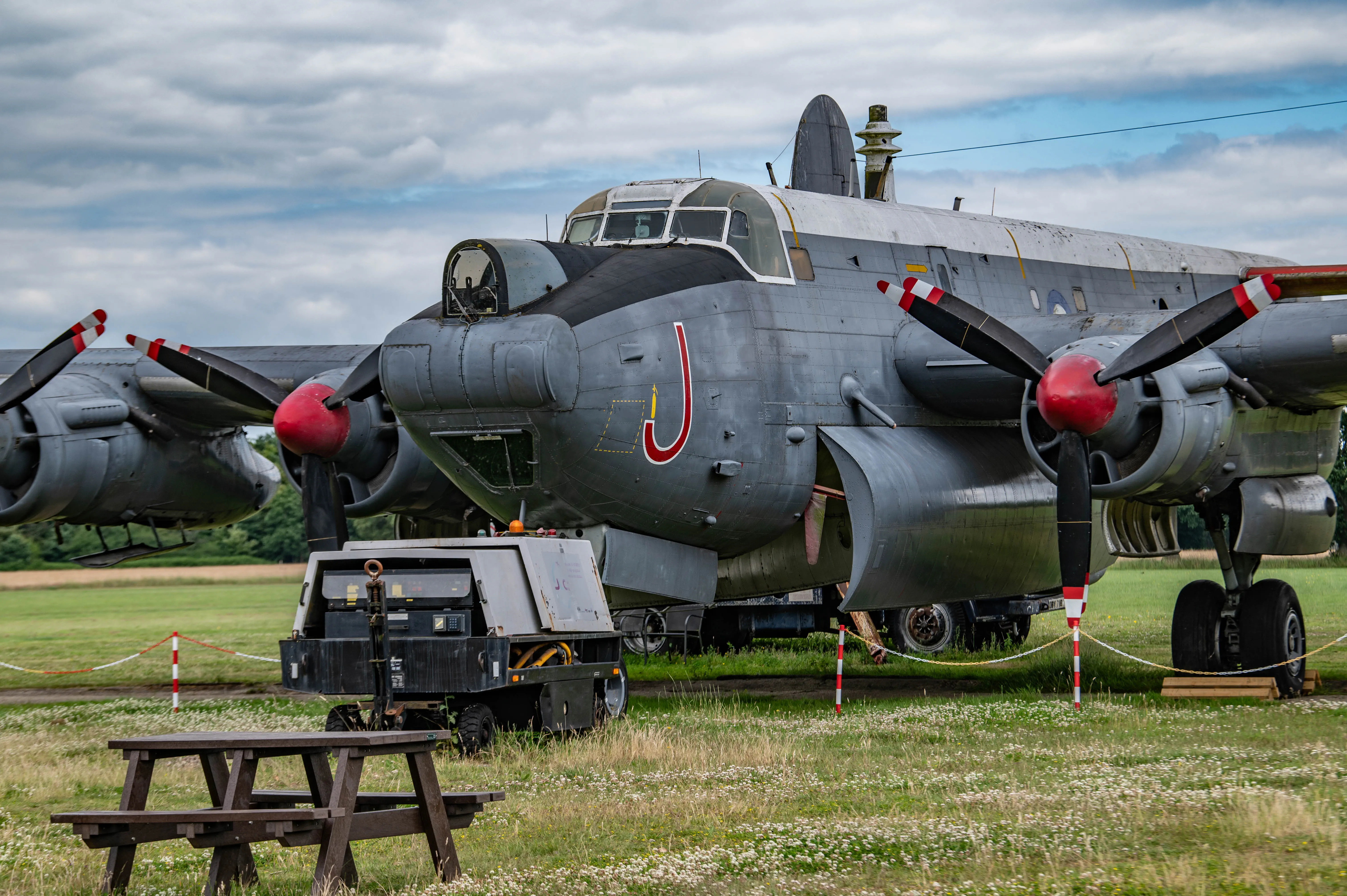 Four-engined vintage military aircraft on a grassy airfield with red-tipped propellers and ground equipment in front.