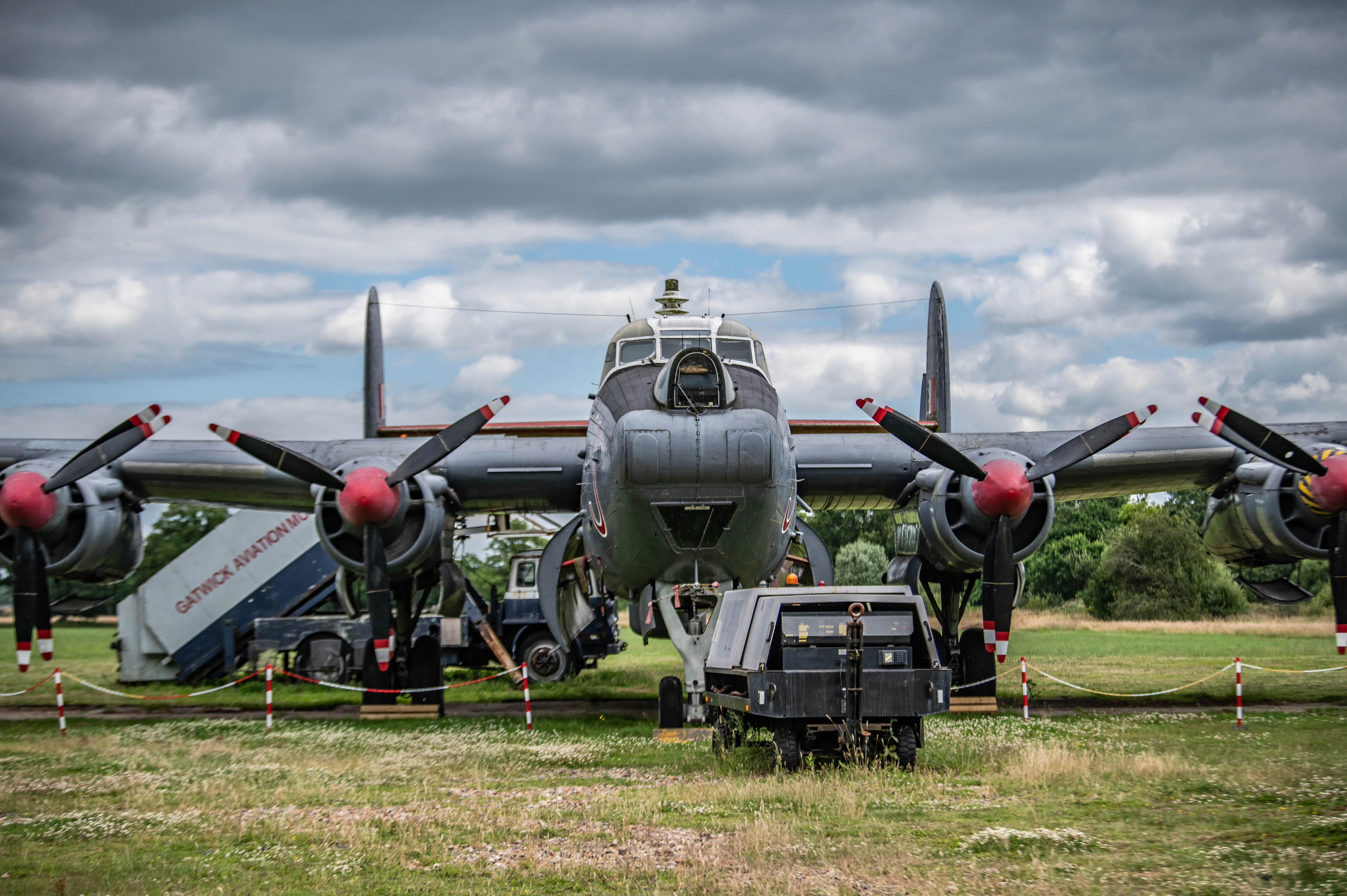 A vintage military aircraft with a gray fuselage and four red-tipped propellers, parked on a grassy field at an aviation museum. The aircraft has a glass nose cone, cockpit windows, and retractable landing gear. In the background, a Gatwick Aviation Museum sign and other aircraft parts are visible, along with a cloudy sky and trees