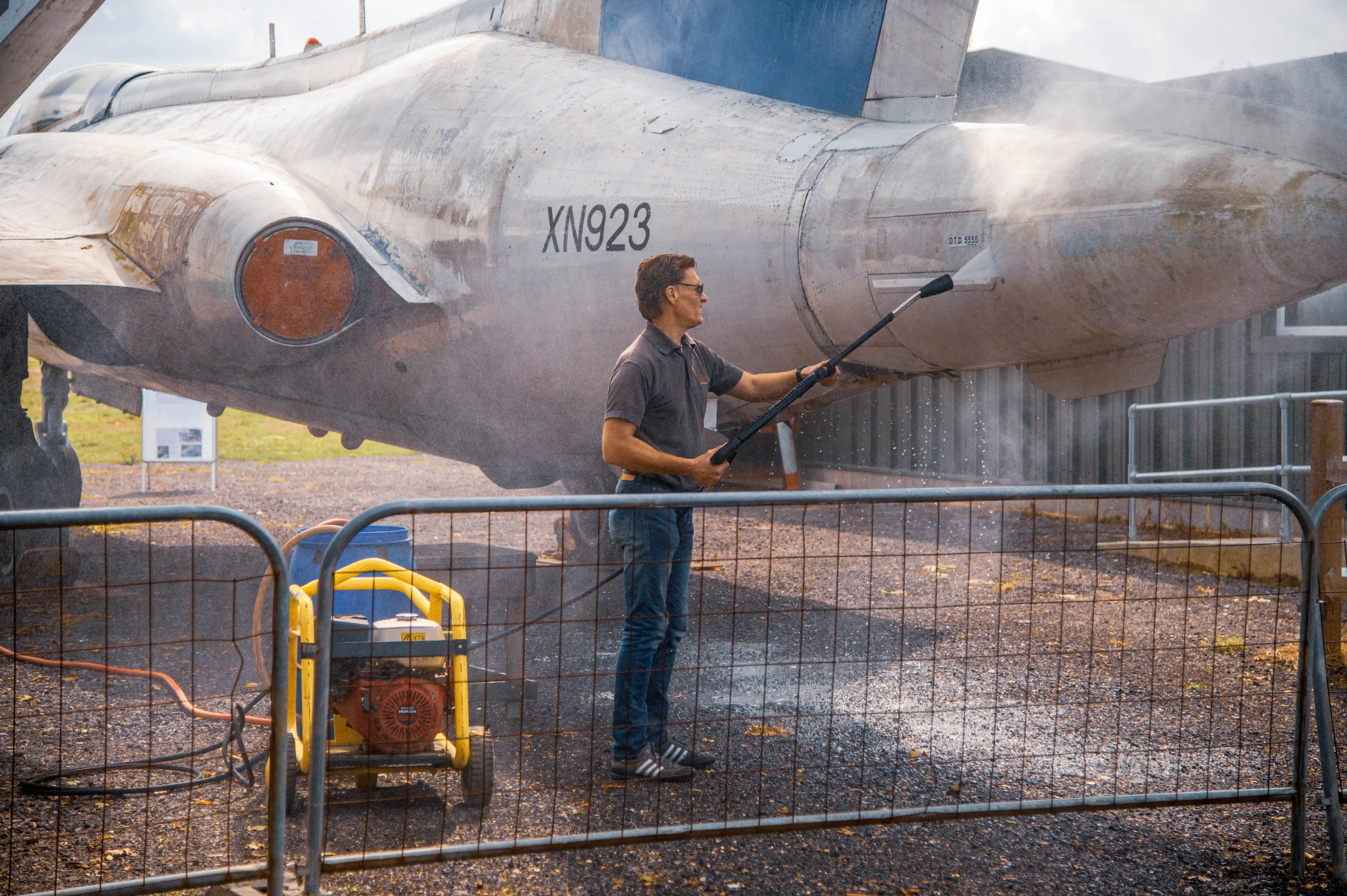 A man in jeans and a grey polo shirt is pressure washing the rear section of a weathered, silver jet aircraft. The aircraft has the registration XN923 visible. A yellow portable generator is nearby, and the scene is cordoned off with a wire fence.