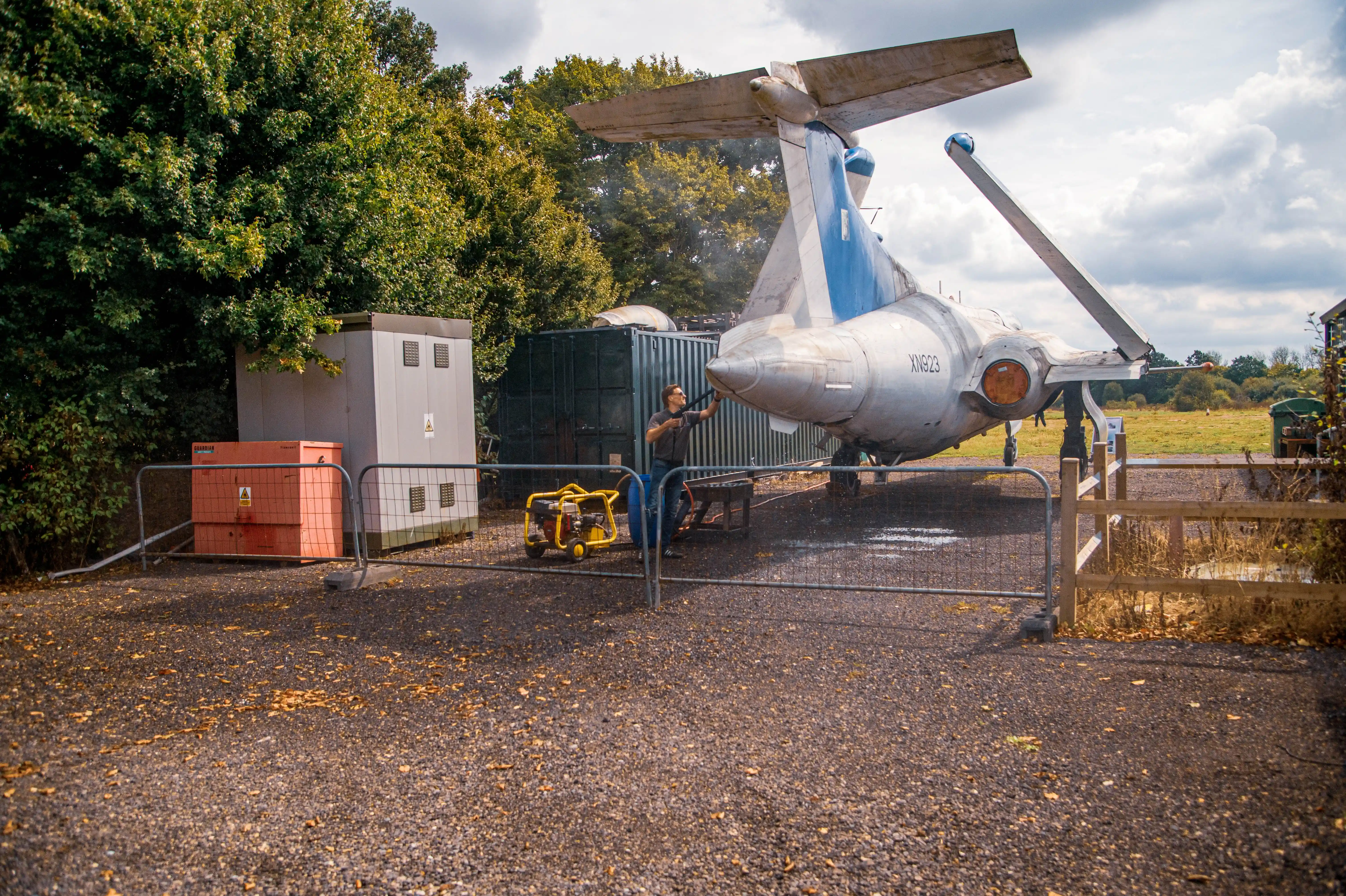 A man is pressure washing the tail section of a silver and blue vintage jet aircraft with registration XN923. He stands behind a metal barrier, holding a pressure washer. Industrial equipment and trees are in the background under a cloudy sky.