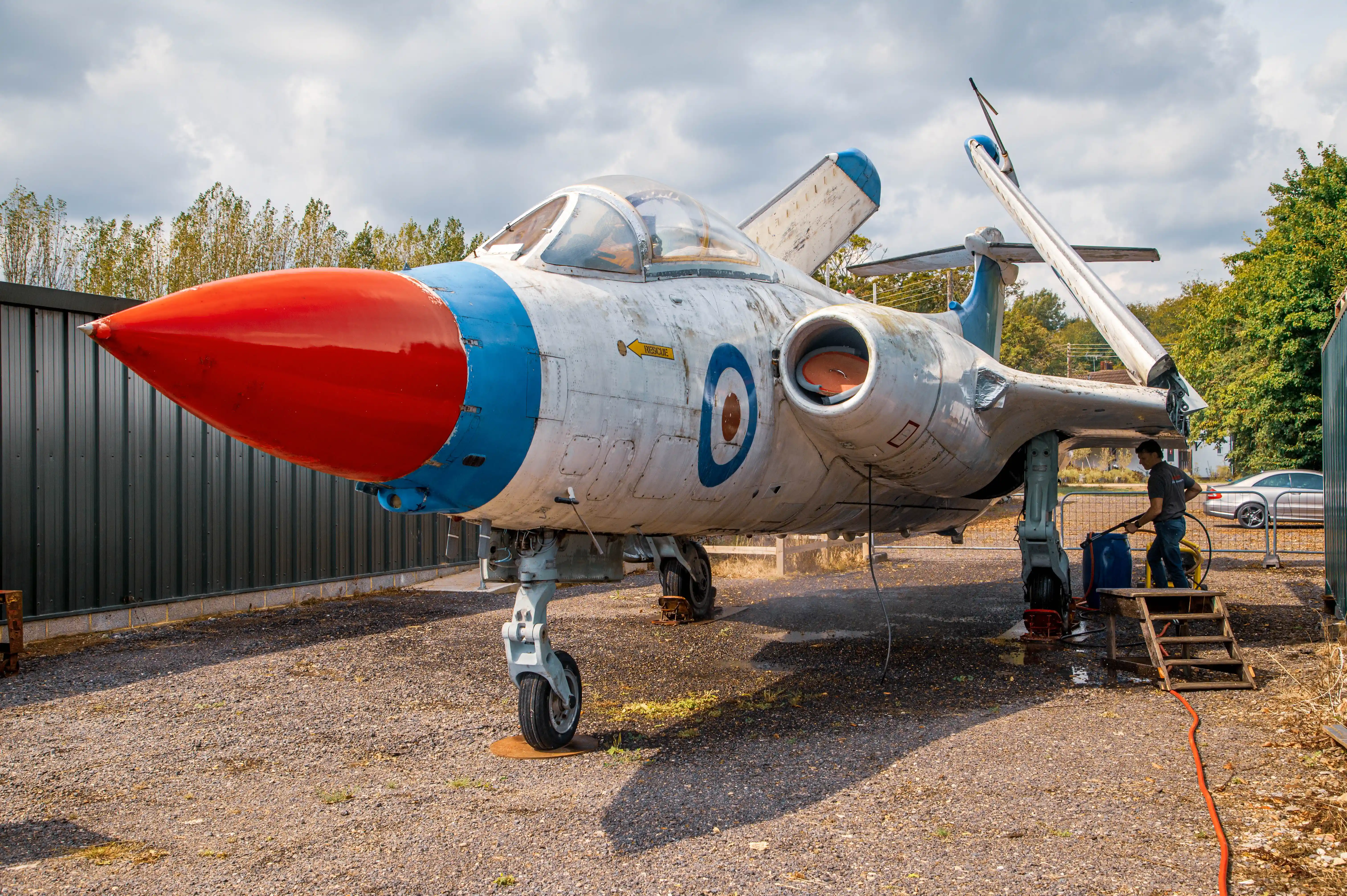 A man washes an old English Electric Lightning jet fighter aircraft which has a red nose cone and blue markings. The plane has the RAF roundel visible.