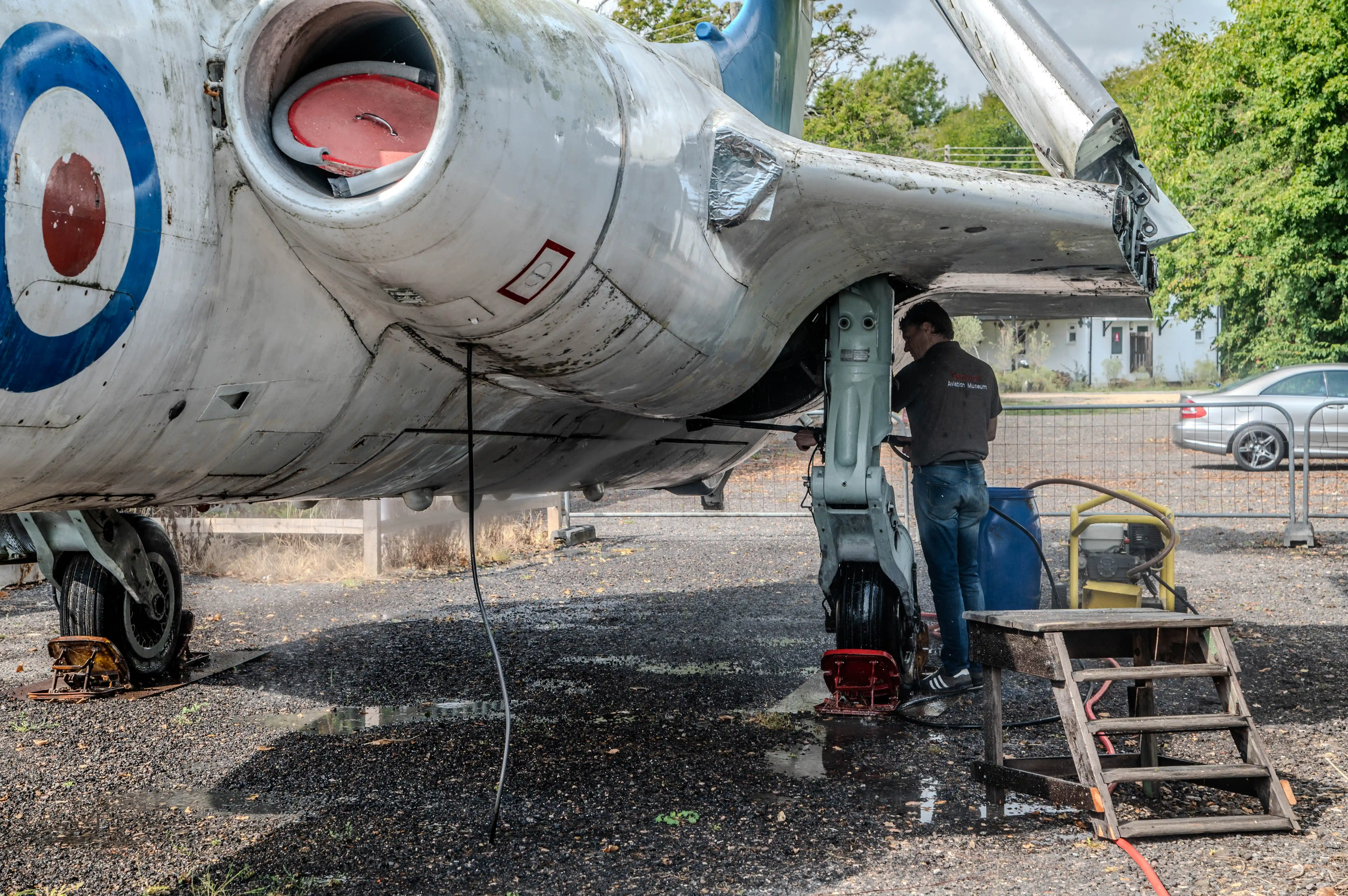A man in a dark shirt and jeans works on the front landing gear of a large, white jet aircraft with a red, white, and blue Royal Air Force roundel visible on its fuselage.
