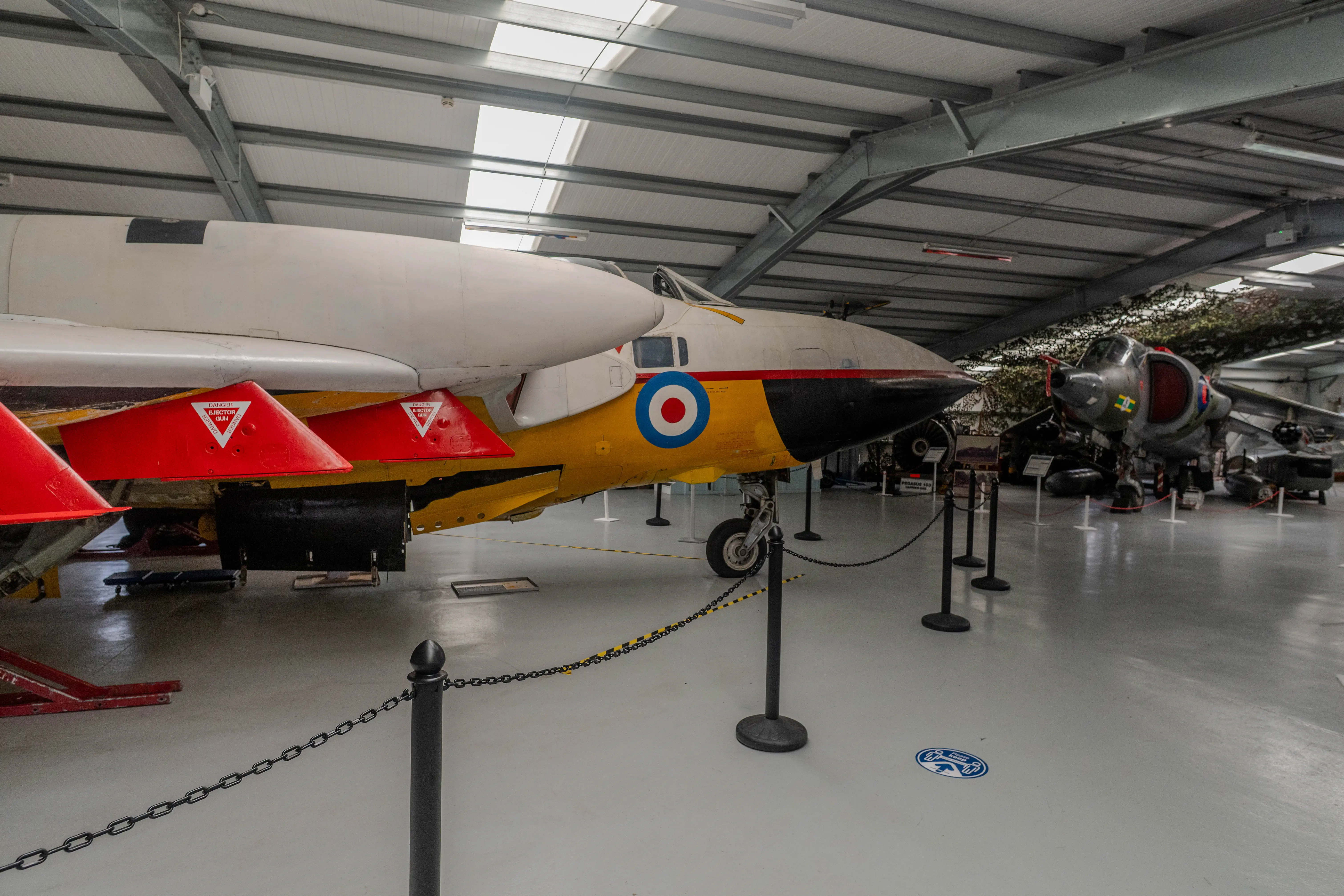 A side view of a vintage British military jet aircraft with a yellow fuselage and white nose, featuring a red and blue roundel on its side. It is parked indoors in a large hangar, with another aircraft visible in the background.