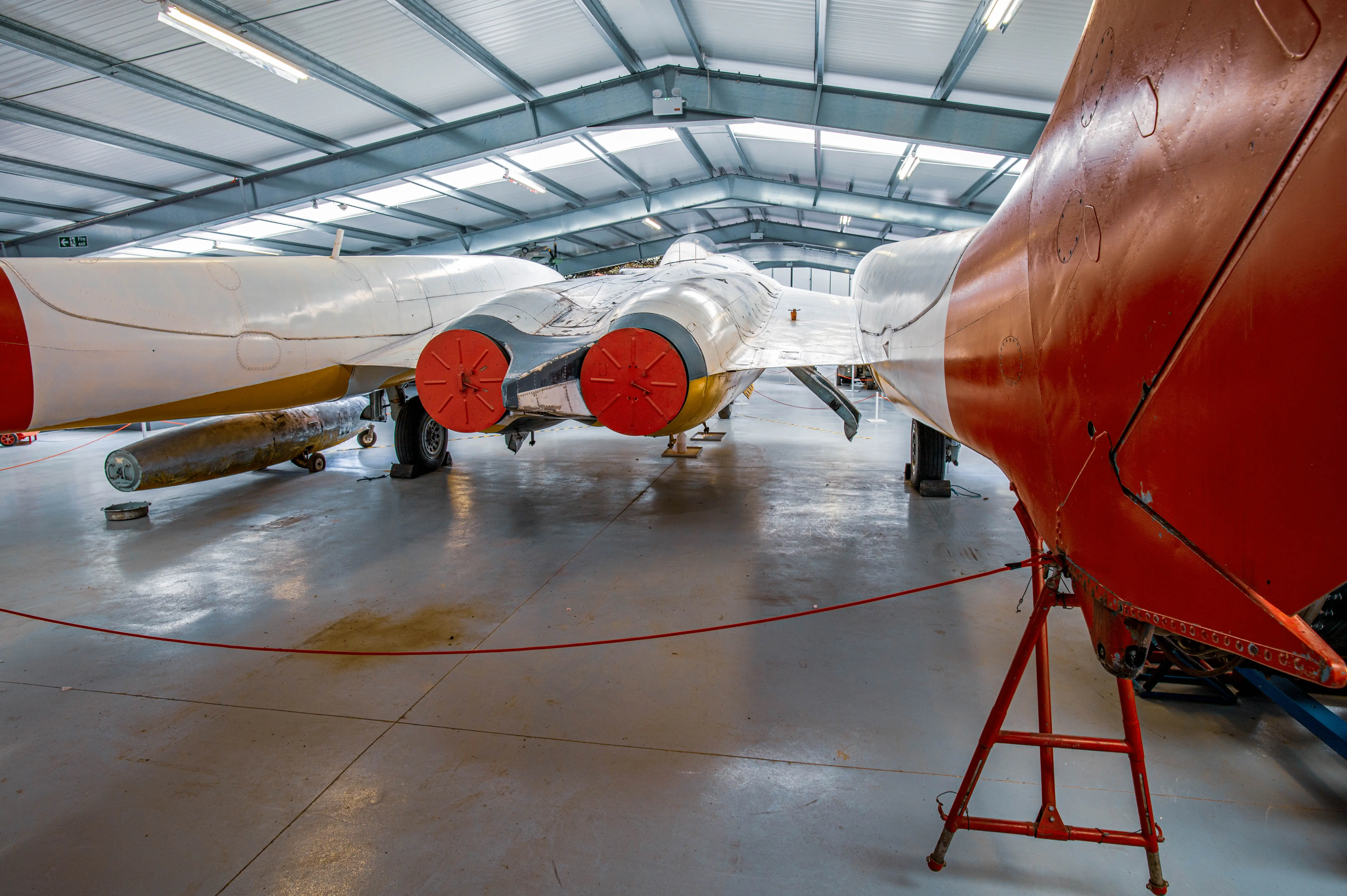 A de Havilland Vampire twin-boom jet aircraft, painted white with red and grey accents, rests on the floor of a large hangar. An unguided rocket pod is mounted beneath its left wing. The aircraft's twin engines, visible at the rear, have bright red intake covers. The metallic roof of the hangar is supported by a series of trusses and illuminated by fluorescent lights.
                              >
                              <figcaption>
                                  <meta itemprop=